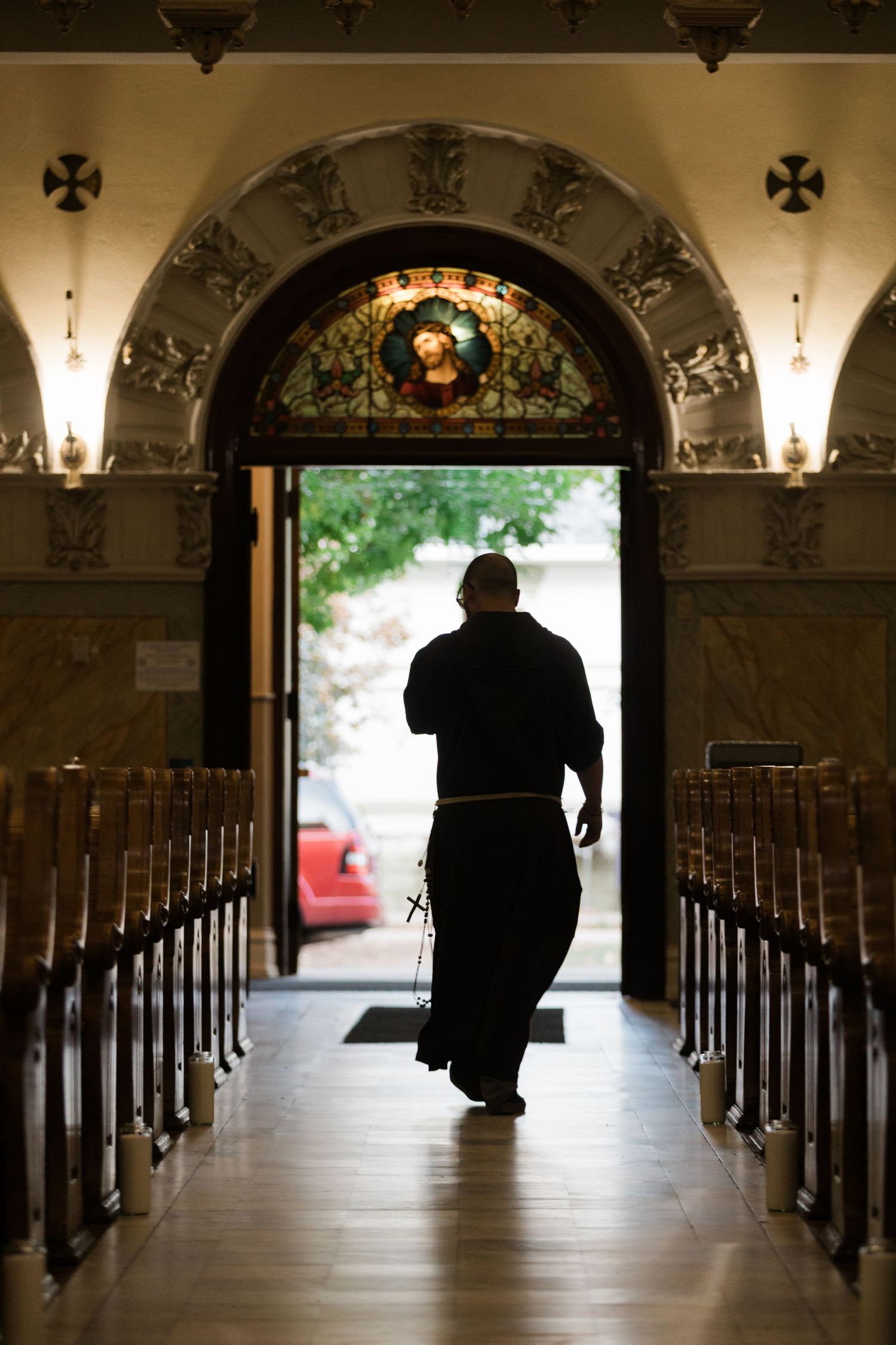 Silhouette of a person walking down church aisle between wooden pews towards bright doorway under ornate arched ceiling.
