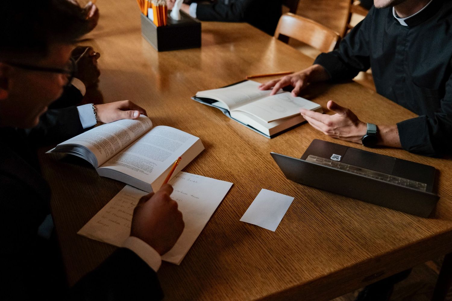 Overhead view of hands writing on papers spread across a wooden table surface in dim lighting.