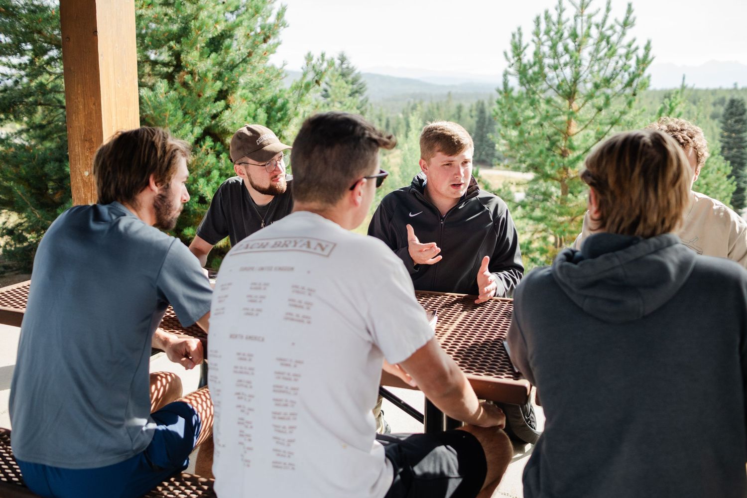 A group of people gathered around a table outdoors with trees visible in the background.