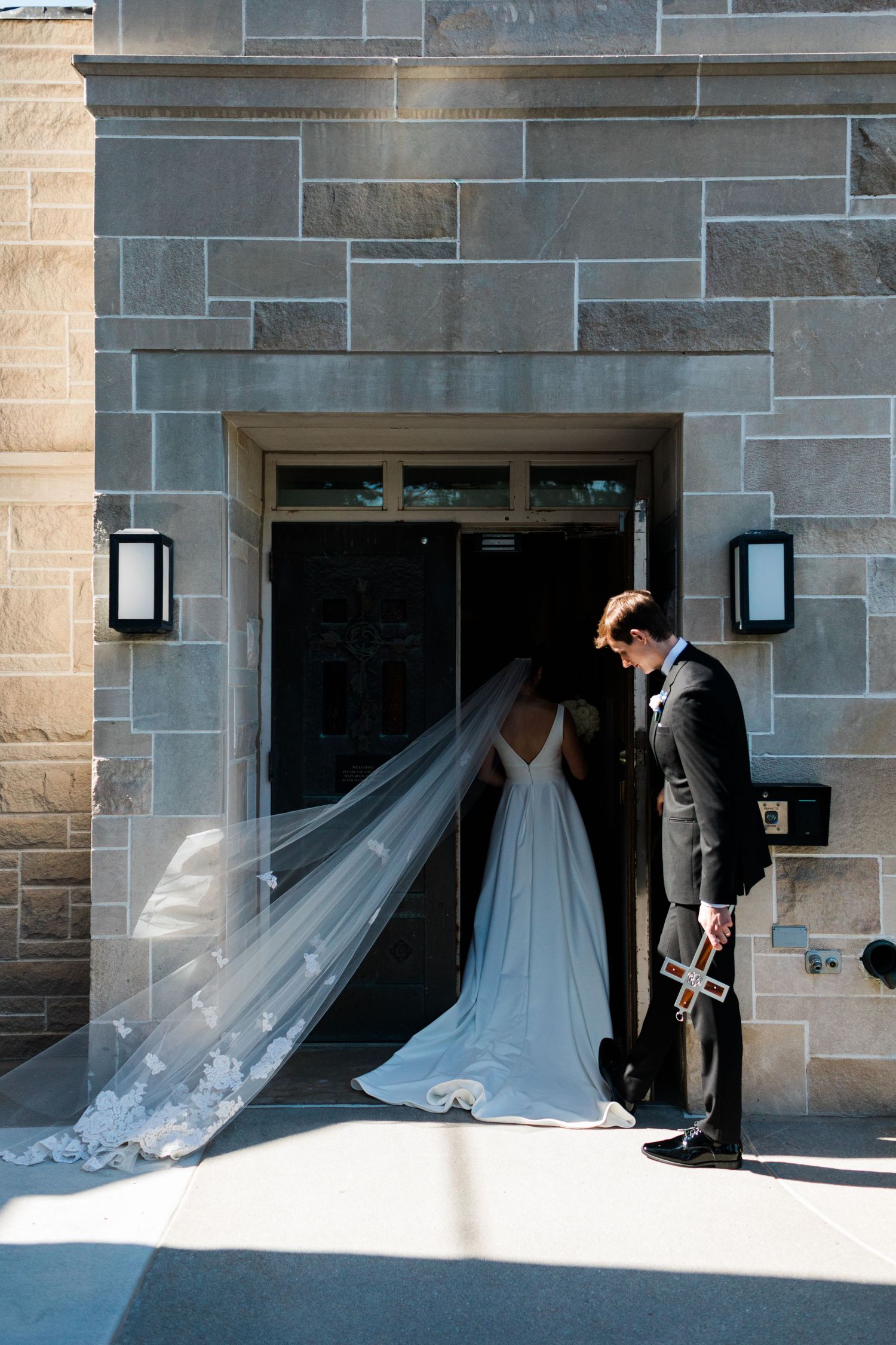 Bride and groom share intimate moment at stone church entrance, her lace veil flowing in the breeze.