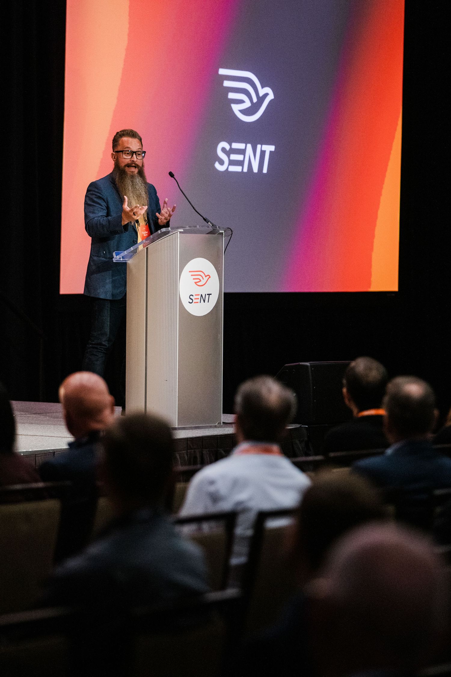 Bearded speaker presents at SENT conference podium on stage with large branded screen backdrop before seated audience.
