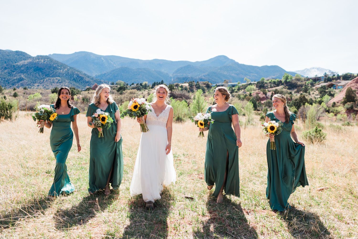 Bride in white gown with bridesmaids in emerald green dresses holding sunflower bouquets in a mountain meadow.