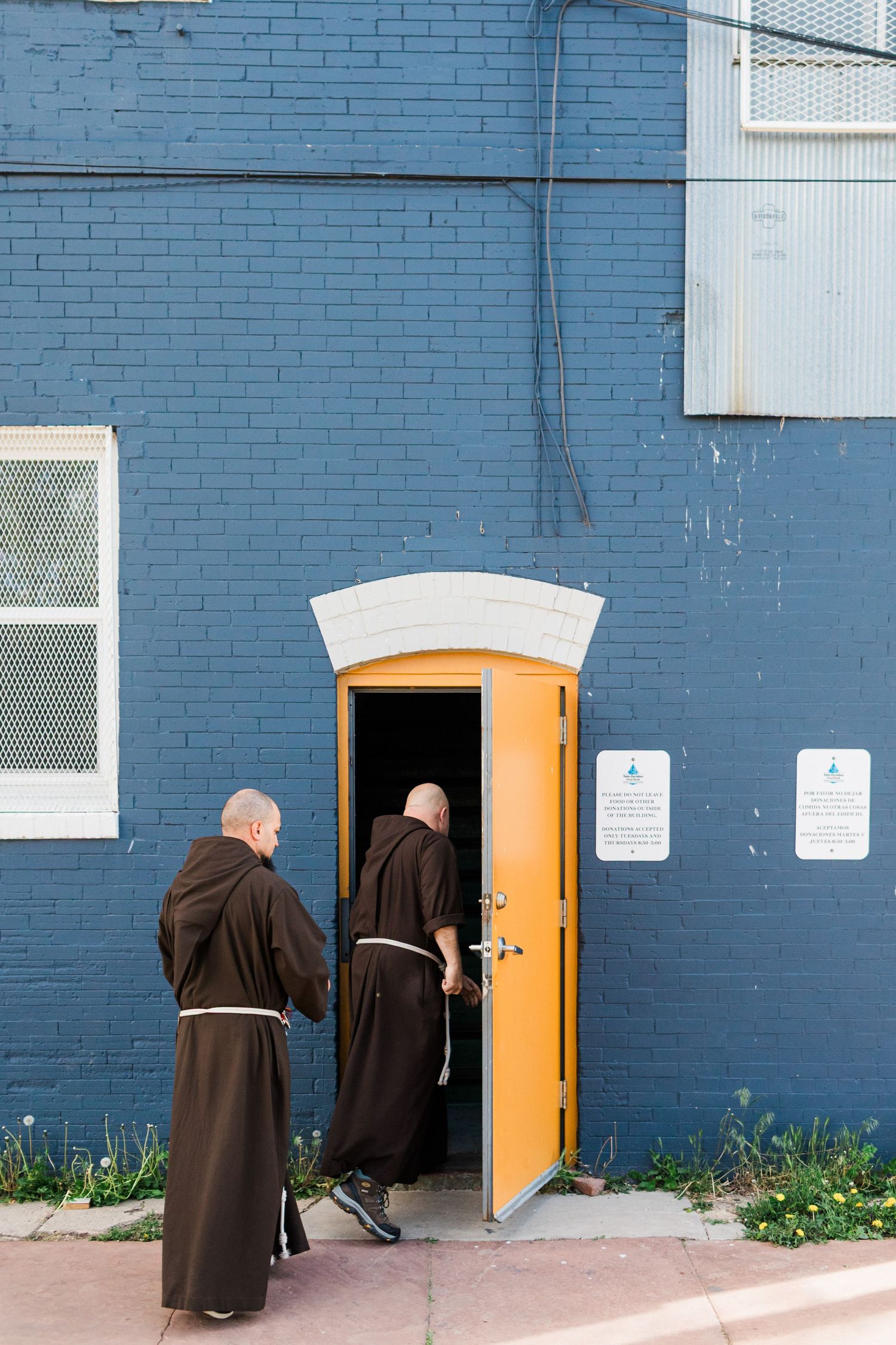Two monks in brown robes entering a yellow arched door on a blue brick building.