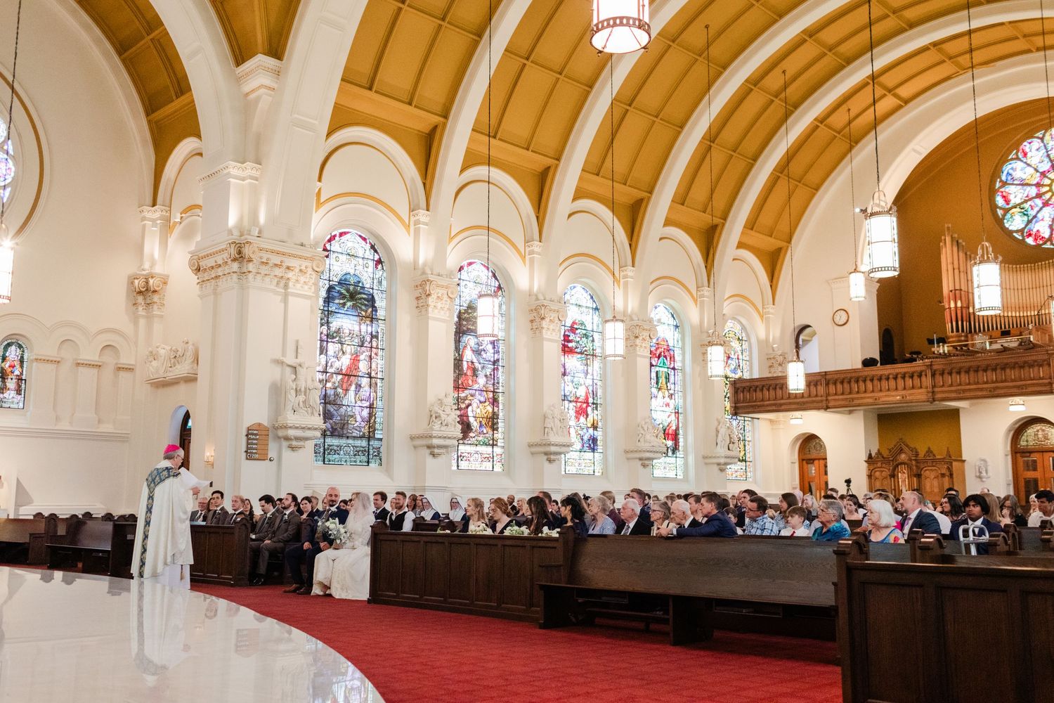 Grand church wedding ceremony with guests seated in pews, stained glass windows, and officiant at altar.