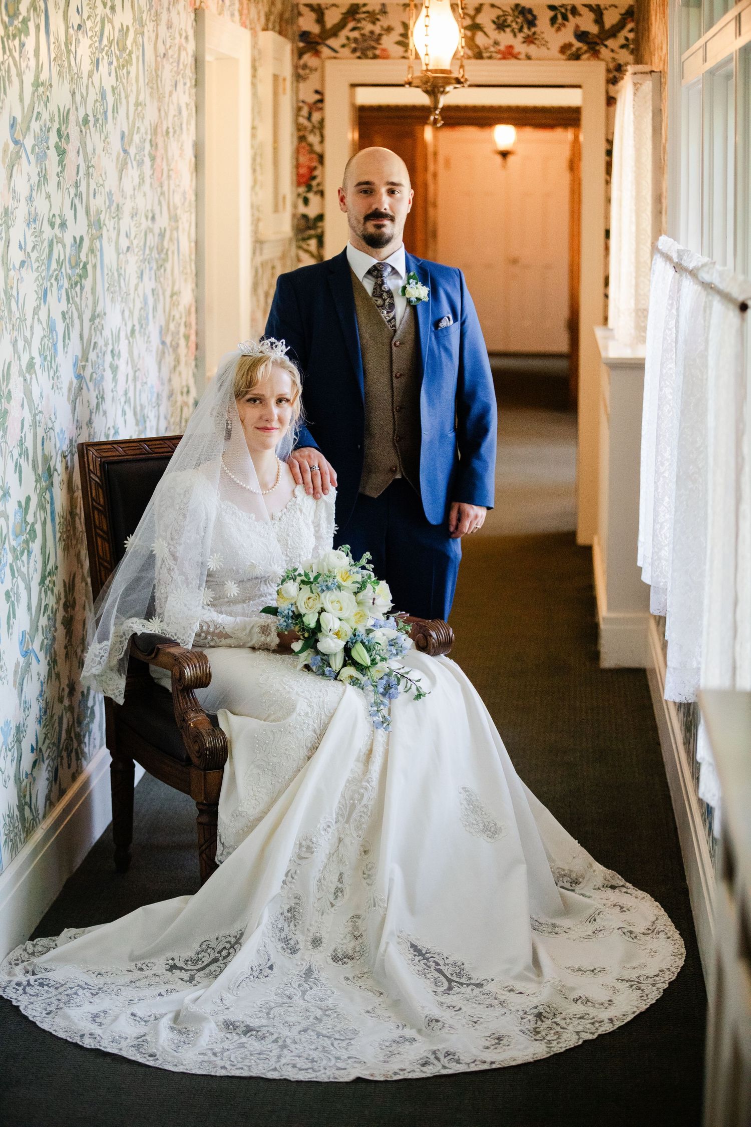Bride in lace gown seated with groom in blue suit posing in floral wallpapered hallway on wedding day.