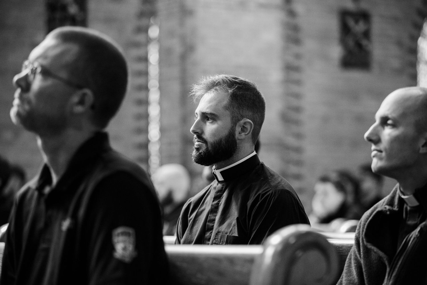 Black and white photo of three men sitting in church pews, middle man in clergy collar gazing forward thoughtfully.
