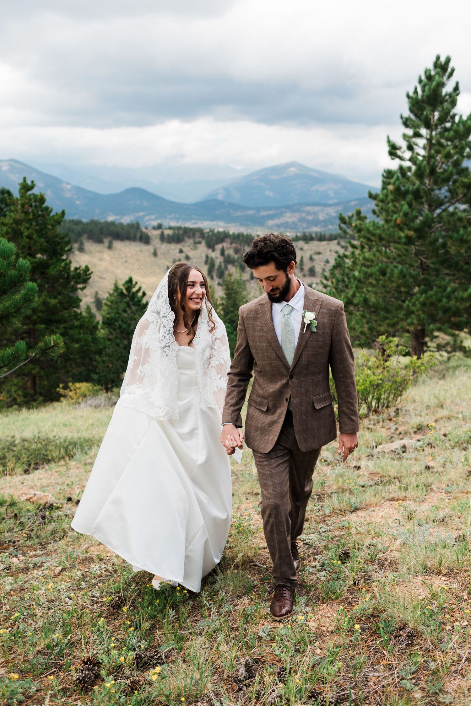 A bride and groom walk hand-in-hand through a mountain meadow, she in a white gown with veil, he in a brown suit.