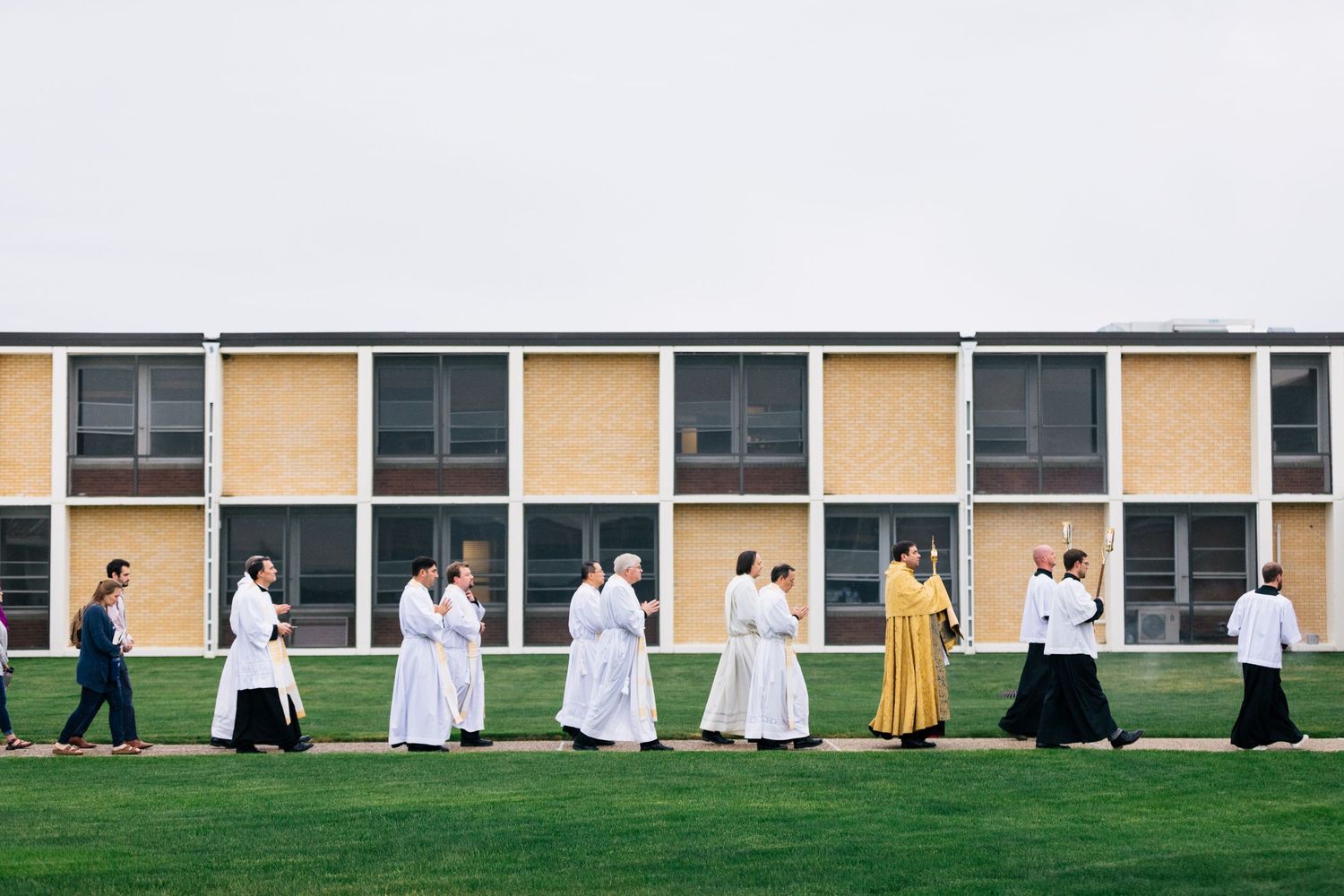 A row of nuns in religious robes walk along a green lawn outside a building.
