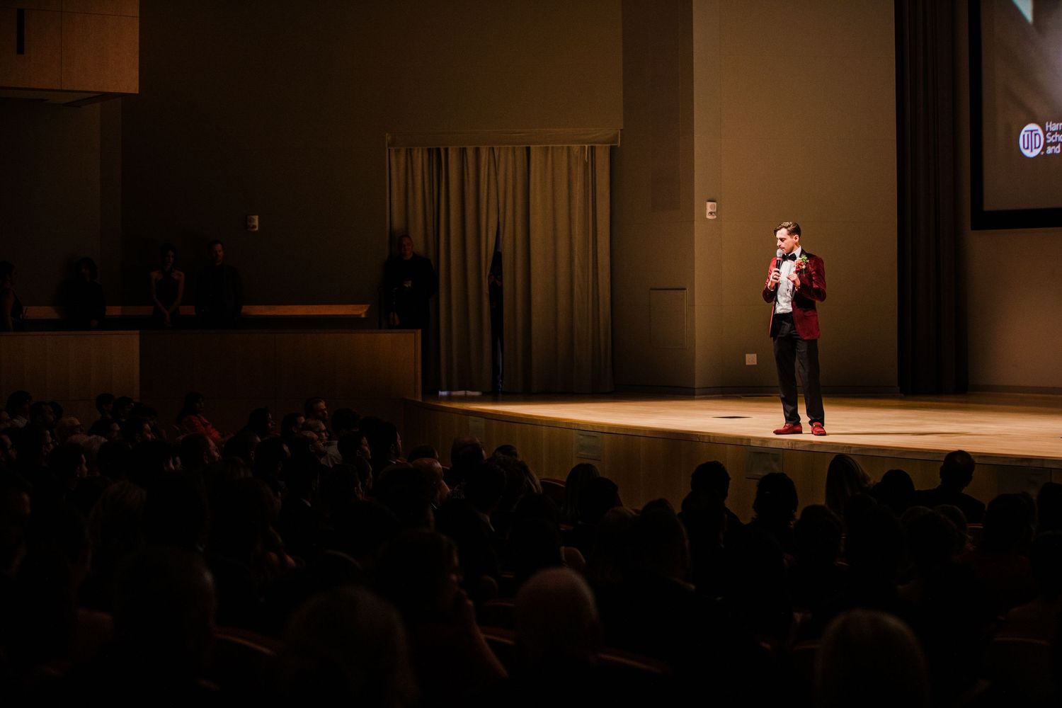 A performer stands on stage in a spotlight during a theatrical performance at an indoor venue.