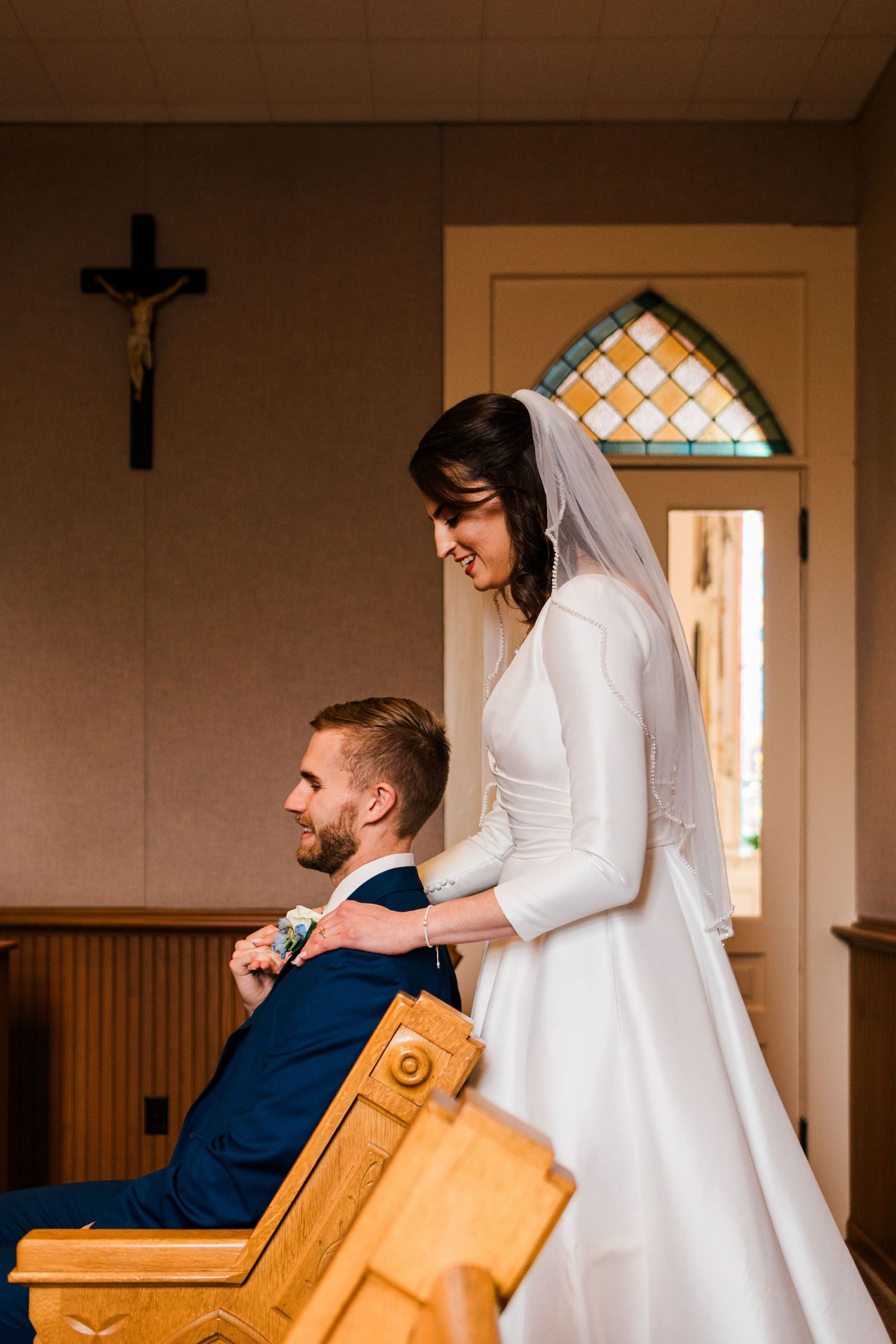 Bride in white gown adjusts groom's boutonniere in a small chapel with crucifix and stained glass window.