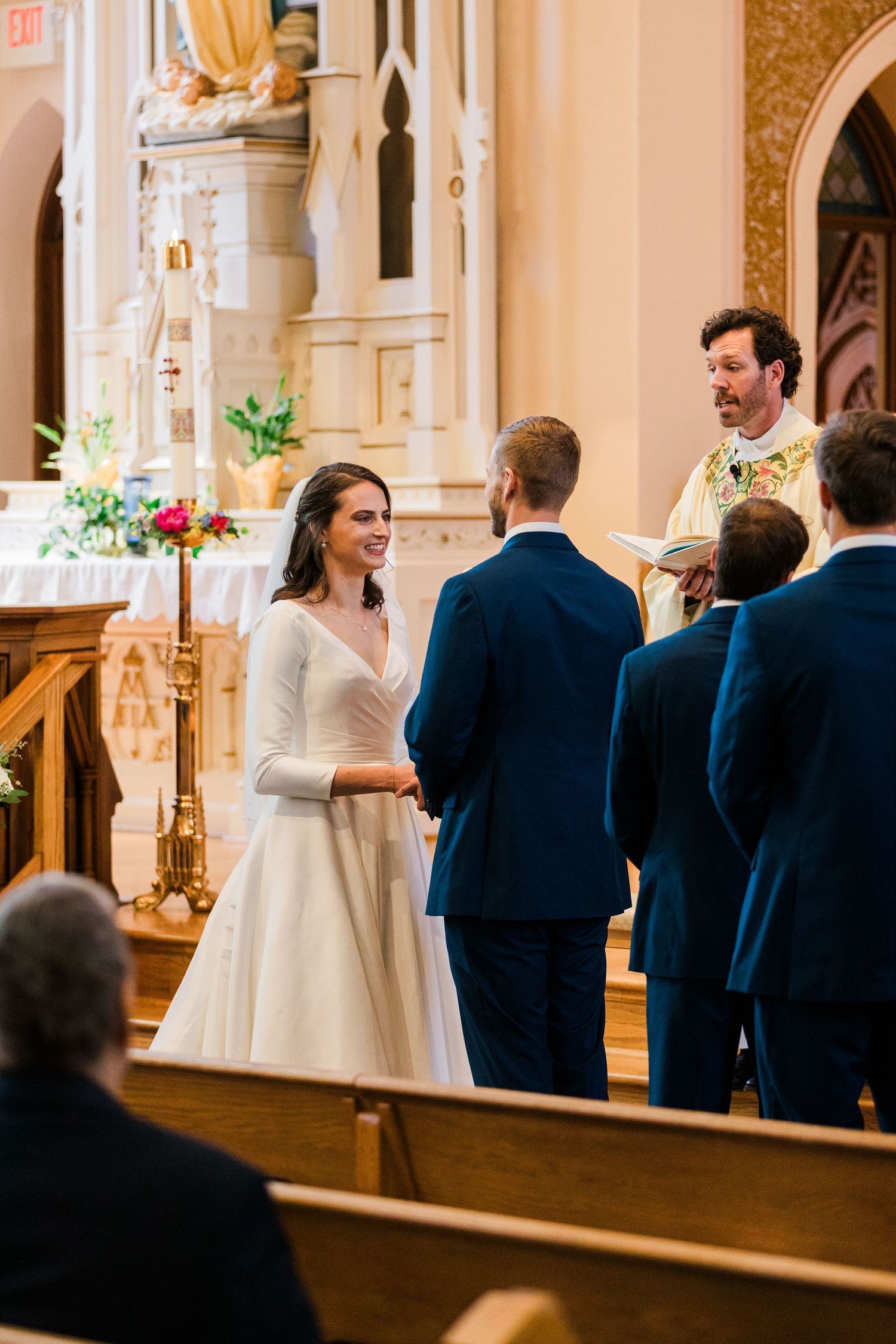 Smiling bride holds groom's hands at church altar during wedding ceremony with priest officiating.