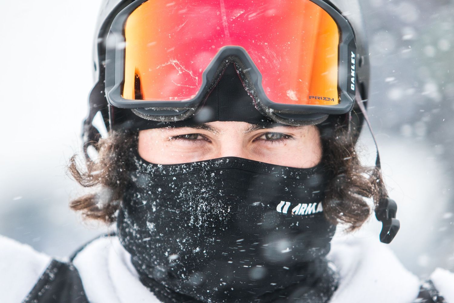 Close up shot of a winter sports enthusiast wearing orange mirrored goggles and black face mask in snowy conditions.