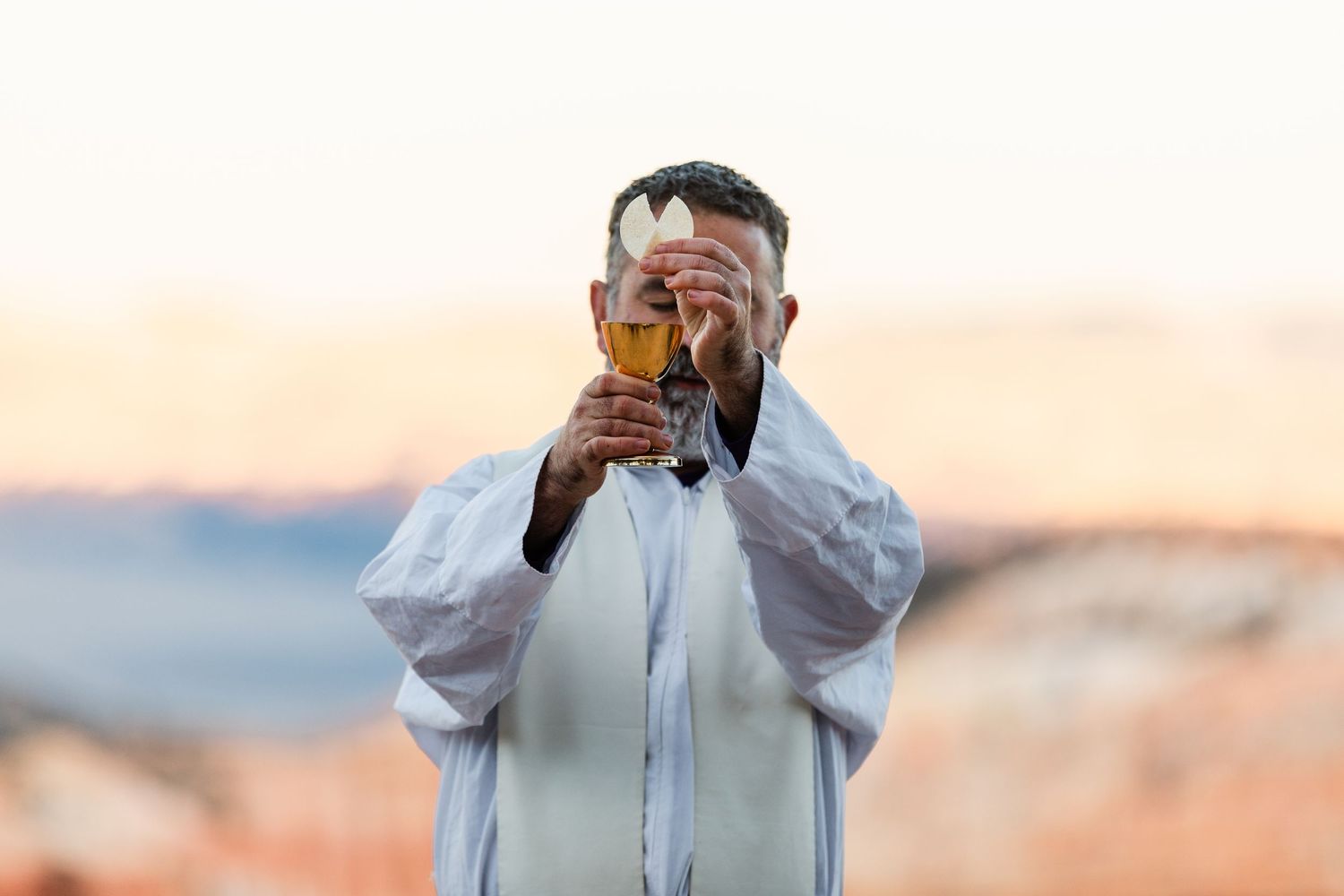 Person in white clothing holds up a glass during sunset against a blurred mountainous background.