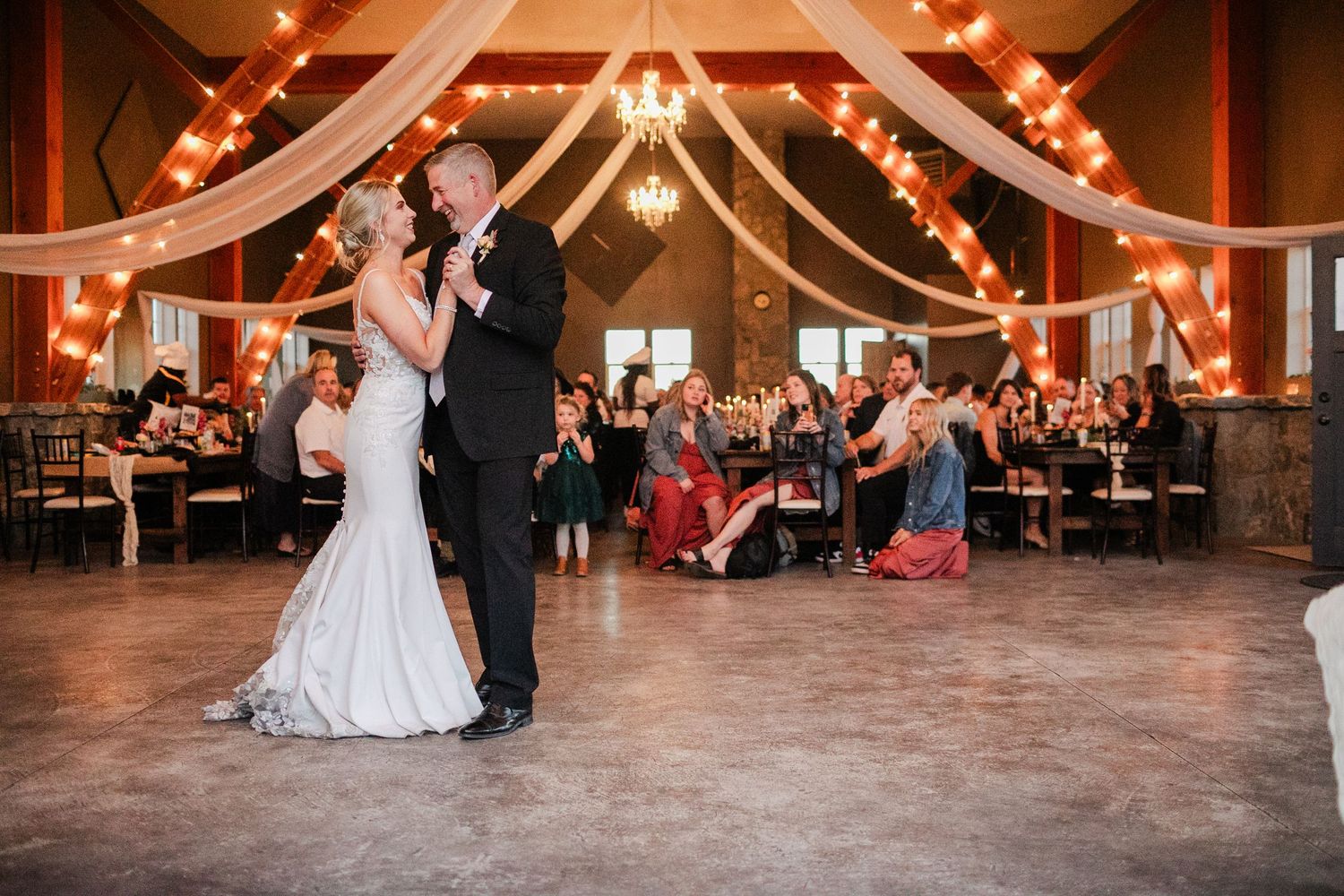 Bride and groom share first dance in elegant barn wedding venue with string lights and draped fabric.