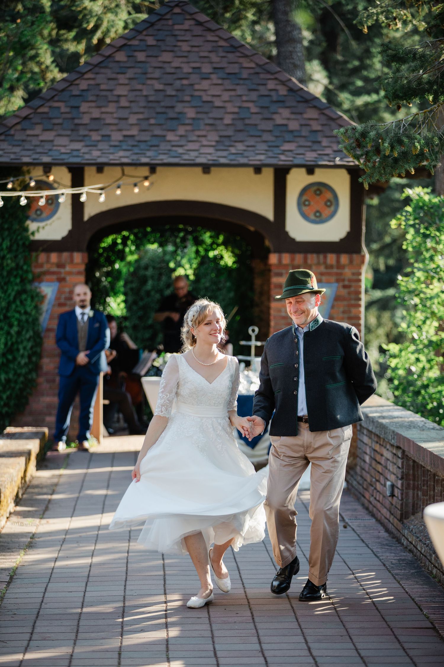 Bride in white dress and groom in traditional attire walk hand-in-hand through a garden archway on their wedding day.