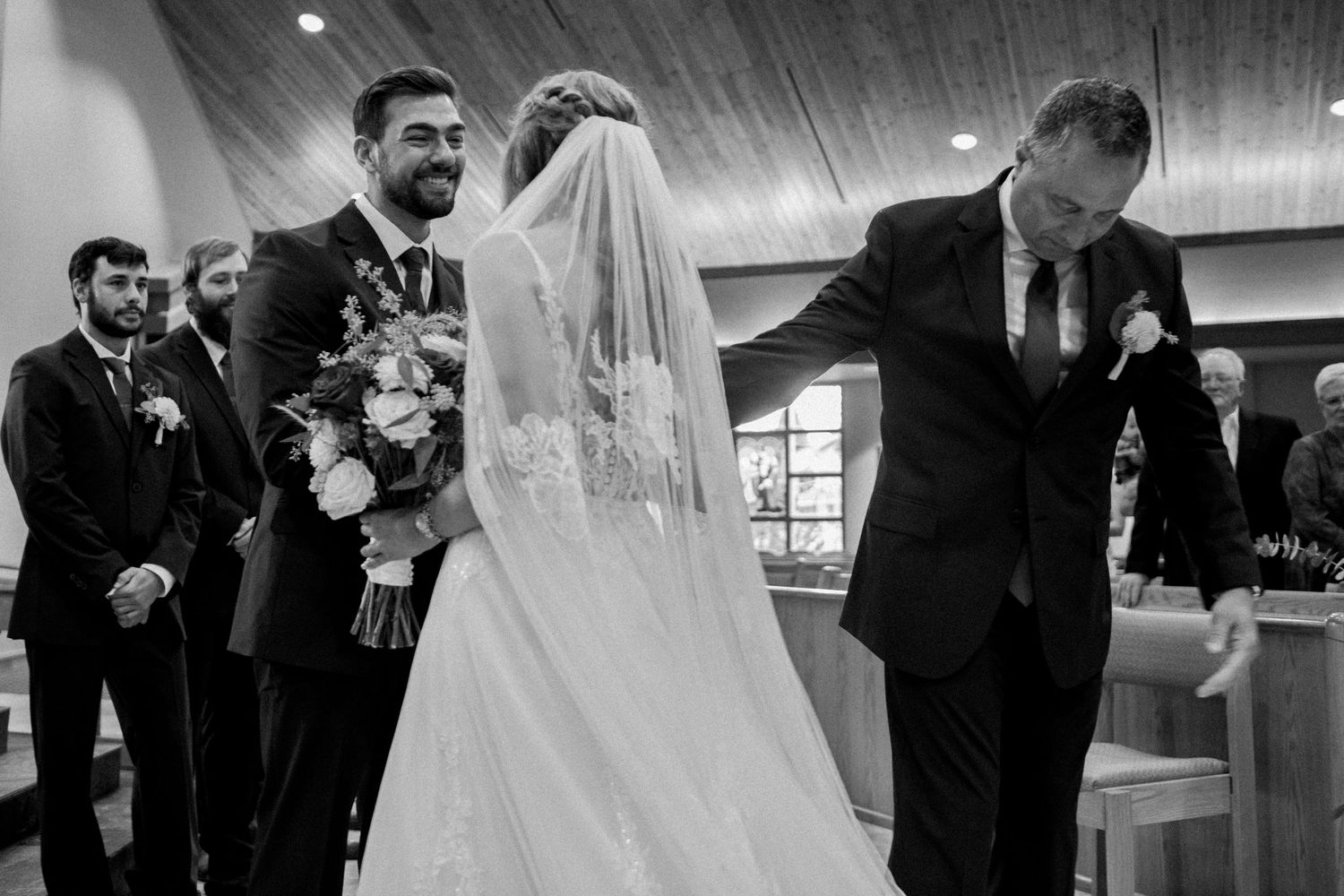 Black and white wedding photo of bride and groom at altar, father bowing emotionally during ceremony.