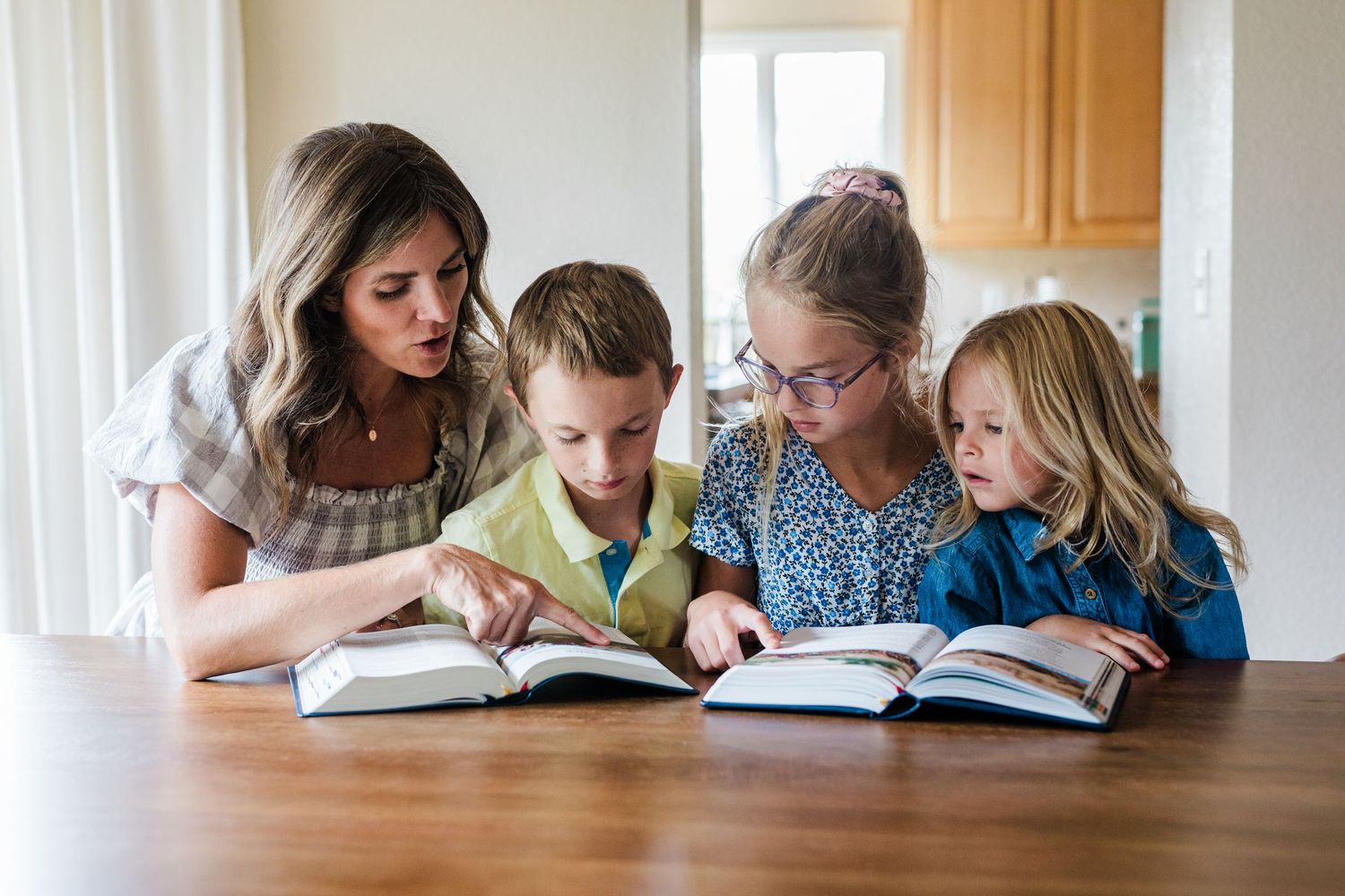 A woman and three children reading an open book together at a wooden table in a bright kitchen.