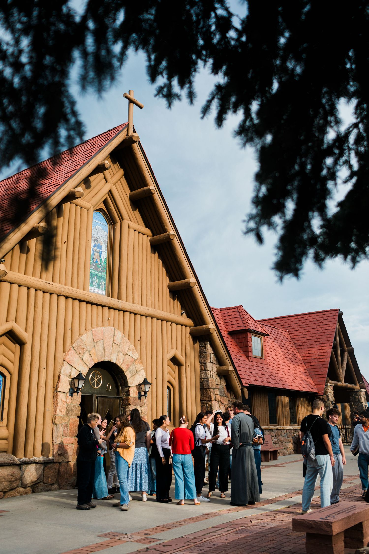 Group of people gathered outside a rustic wooden church with a cross, red roof, and stained glass window under a blue sky.