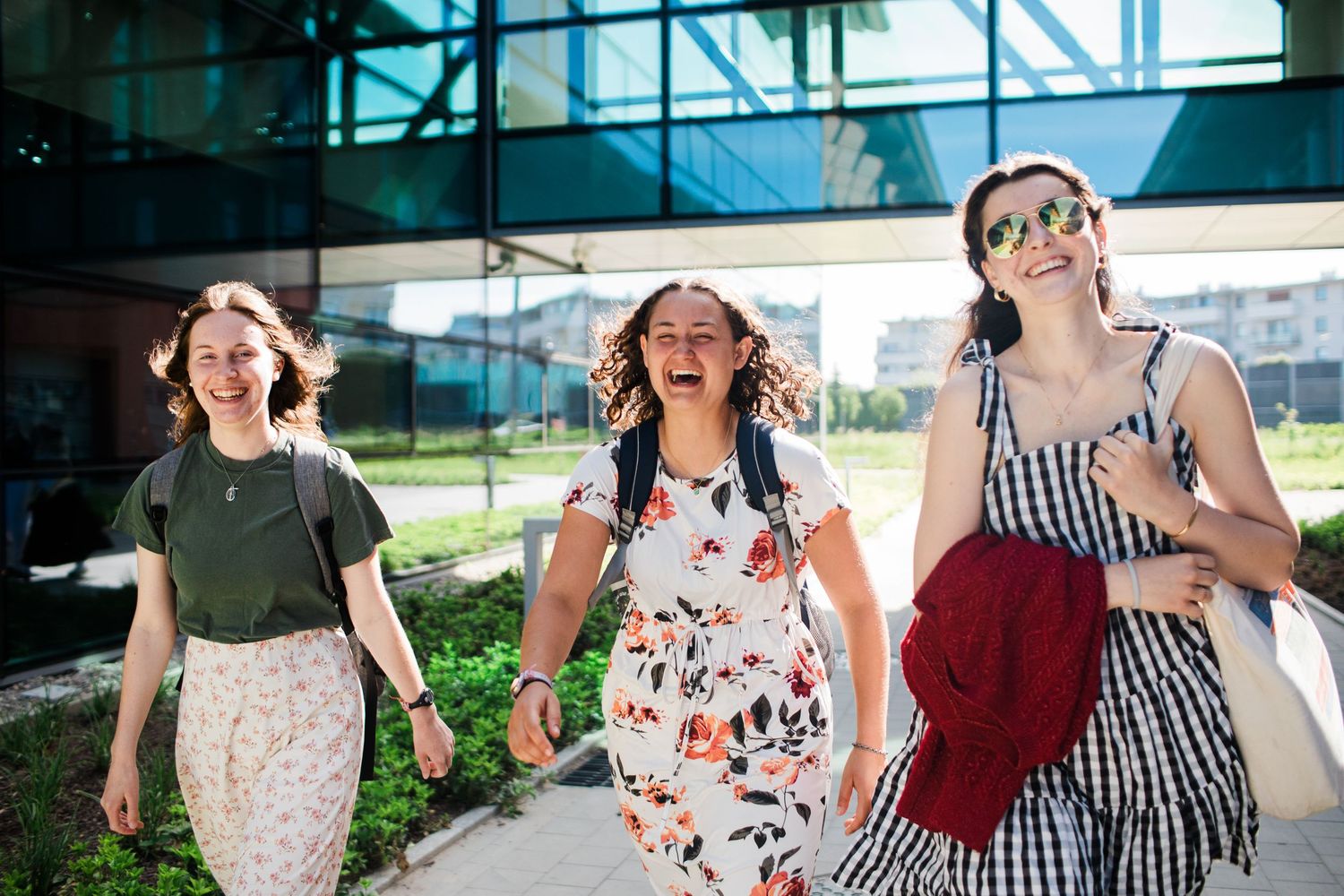 Three young women laughing and walking together outdoors near a modern glass building on a sunny day.