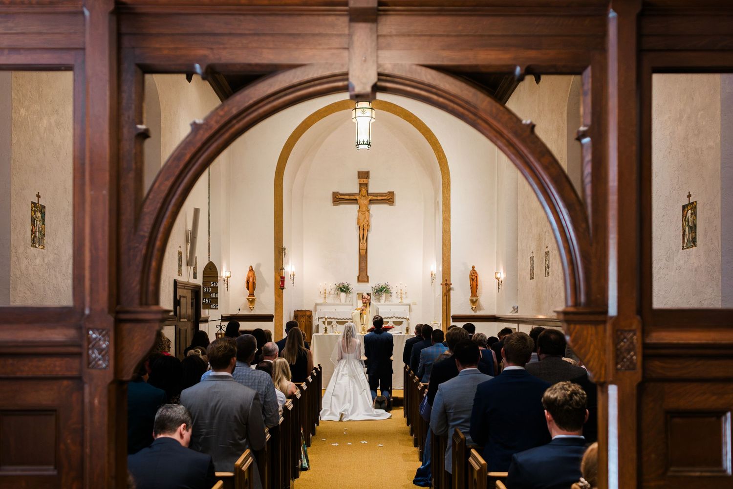Catholic church wedding ceremony with bride and groom at altar beneath cross, guests seated in pews, viewed through wooden arch.