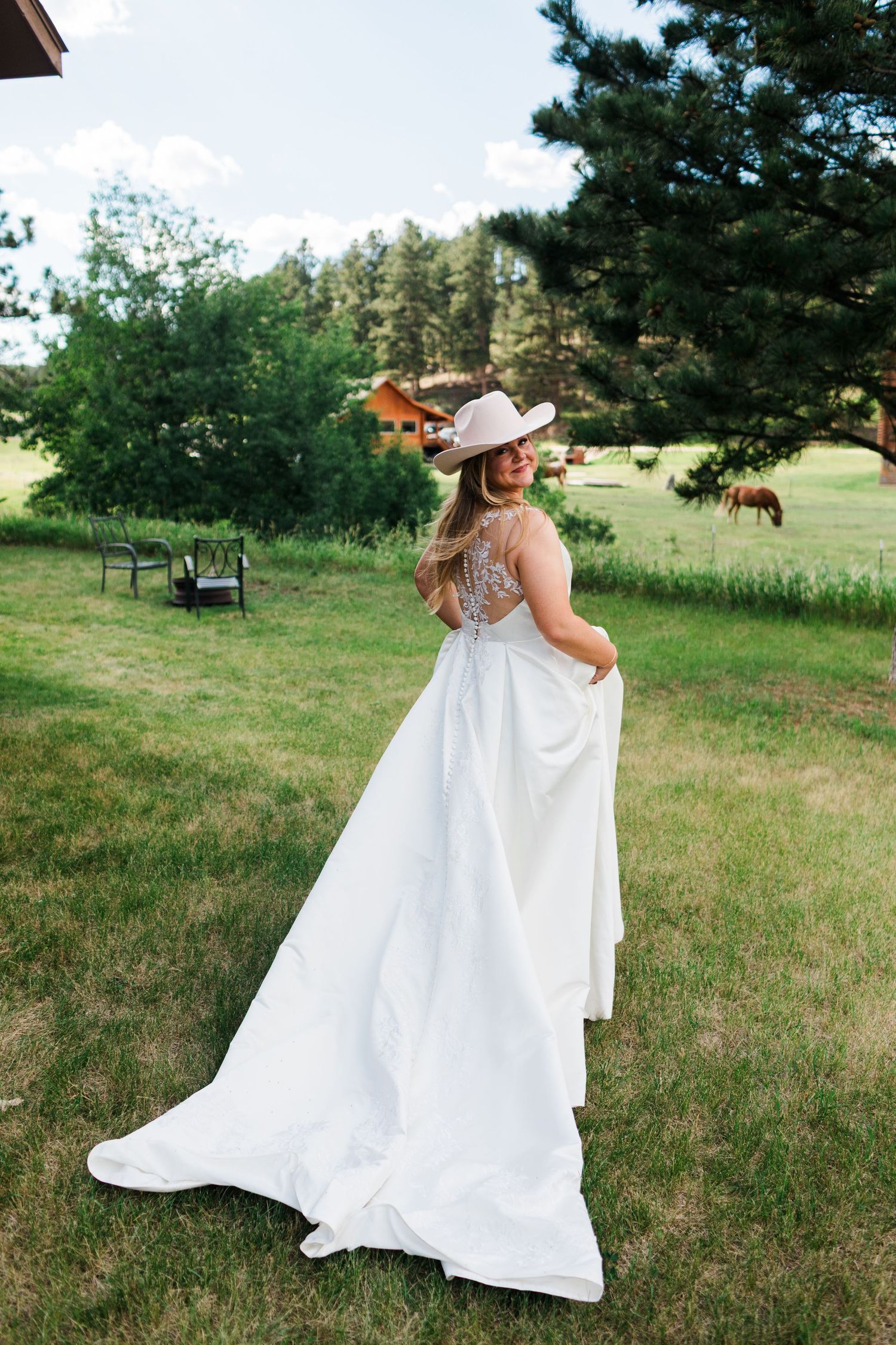 Bride in white wedding gown and pink cowboy hat twirling on green lawn with horses and barn in background.