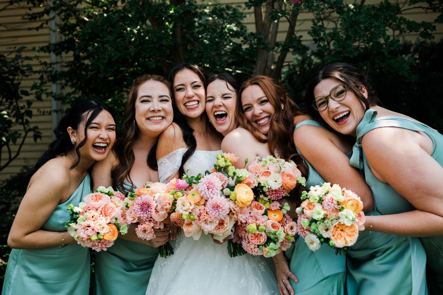 Bride and bridesmaids in mint dresses laughing and holding colorful bouquets outdoors.