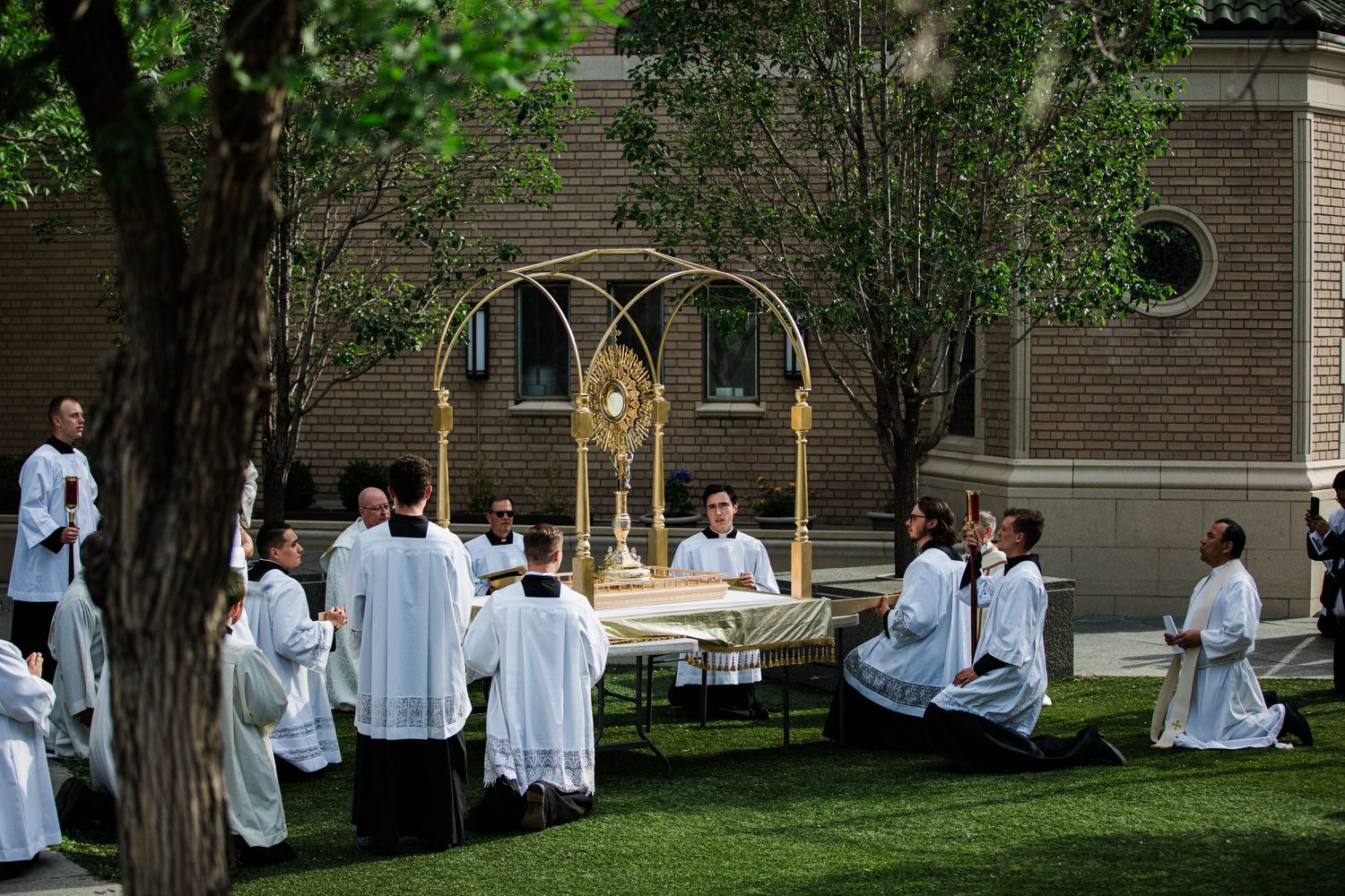Catholic clergy and altar servers in white robes kneel outdoors during a Eucharistic procession with golden monstrance.