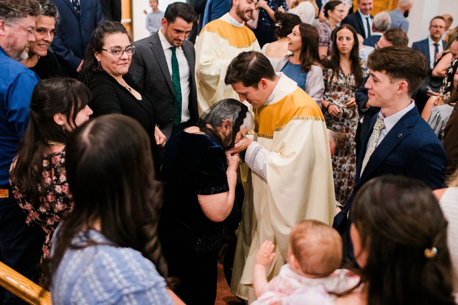 Priest blessing a kneeling woman during a Catholic ceremony, surrounded by family members in a church.