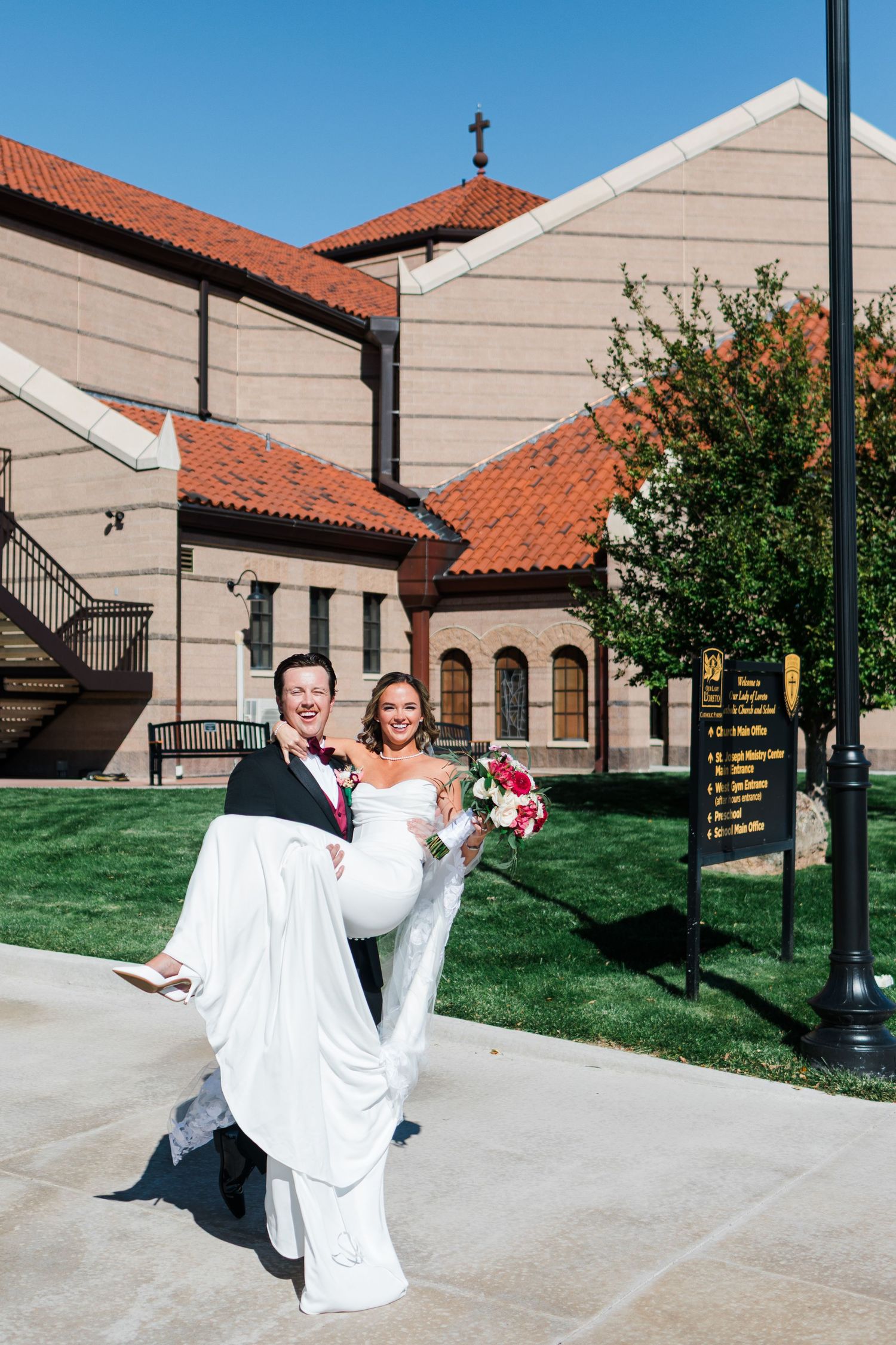 Groom carries bride in white gown outside a church with red tile roof on their wedding day.