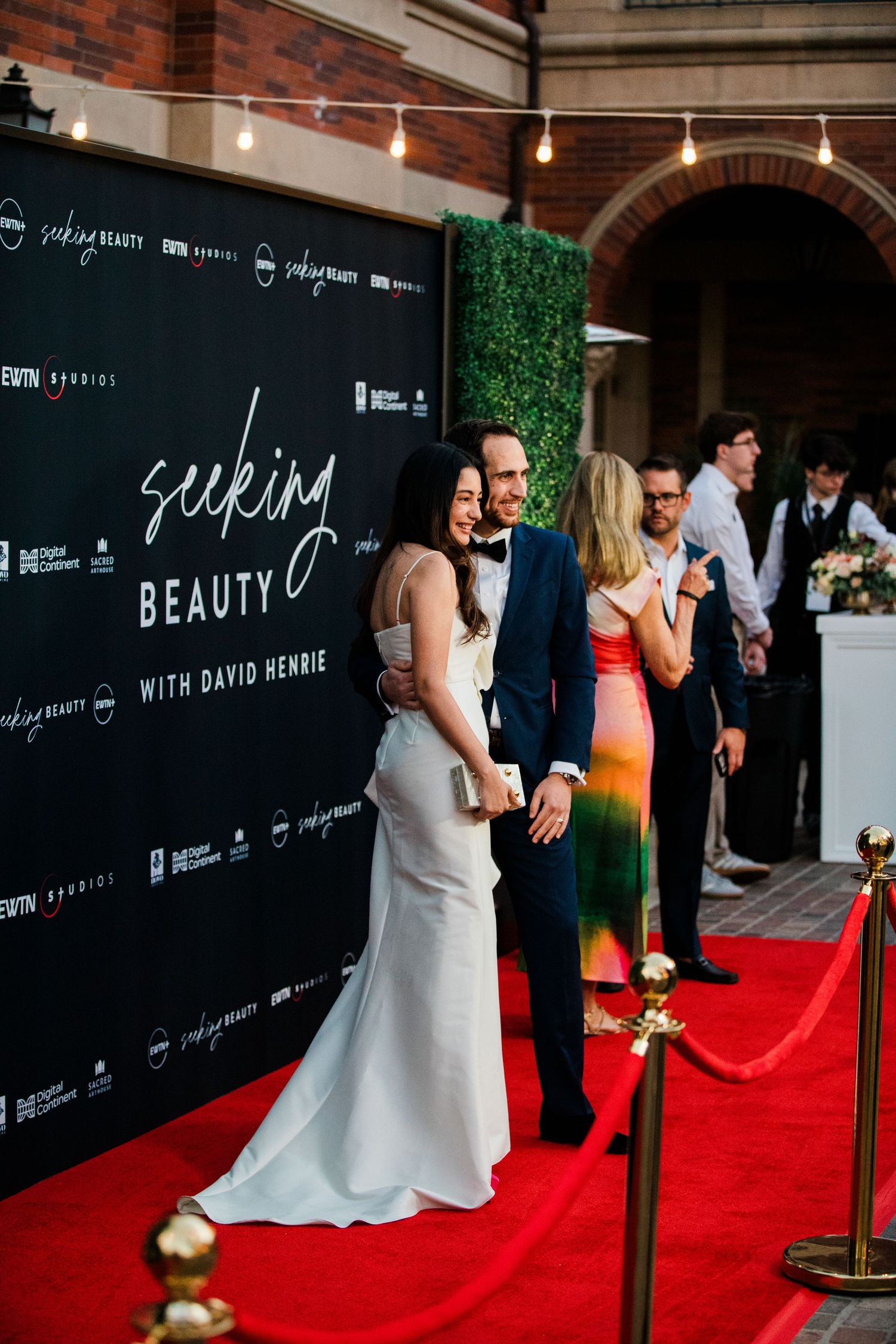 Guests pose on a red carpet at a formal event with black backdrop and lighting setup.