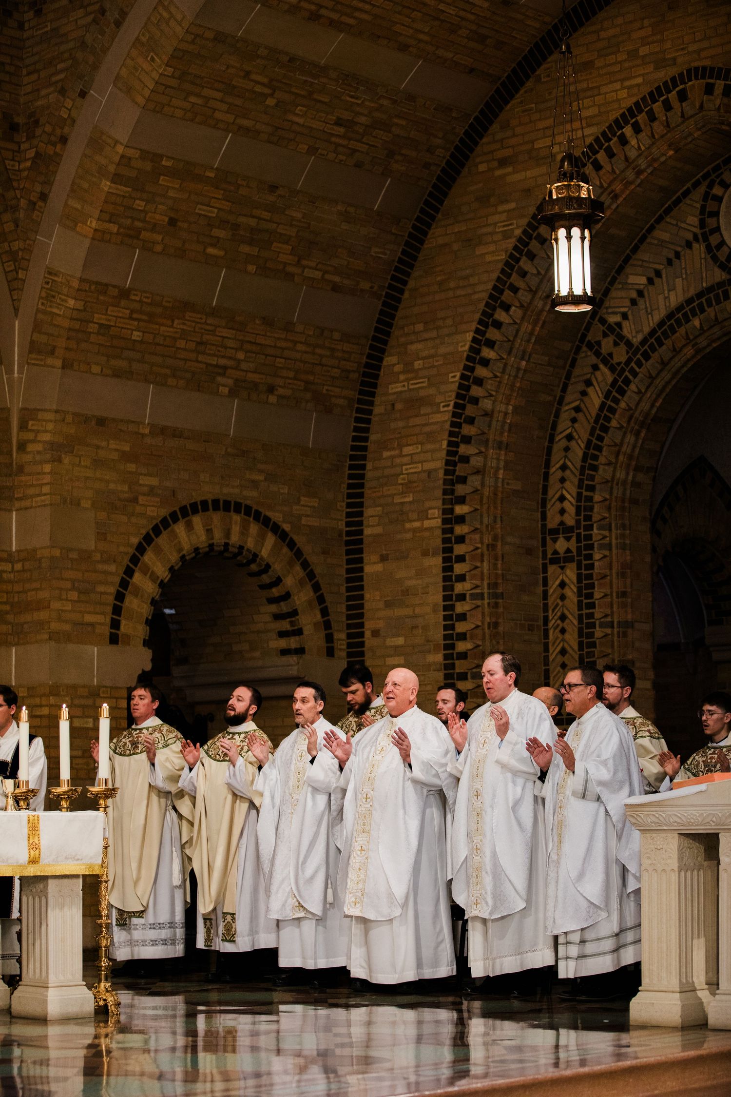 Catholic priests in white vestments stand at the altar during a Mass in a historic brick arched church.