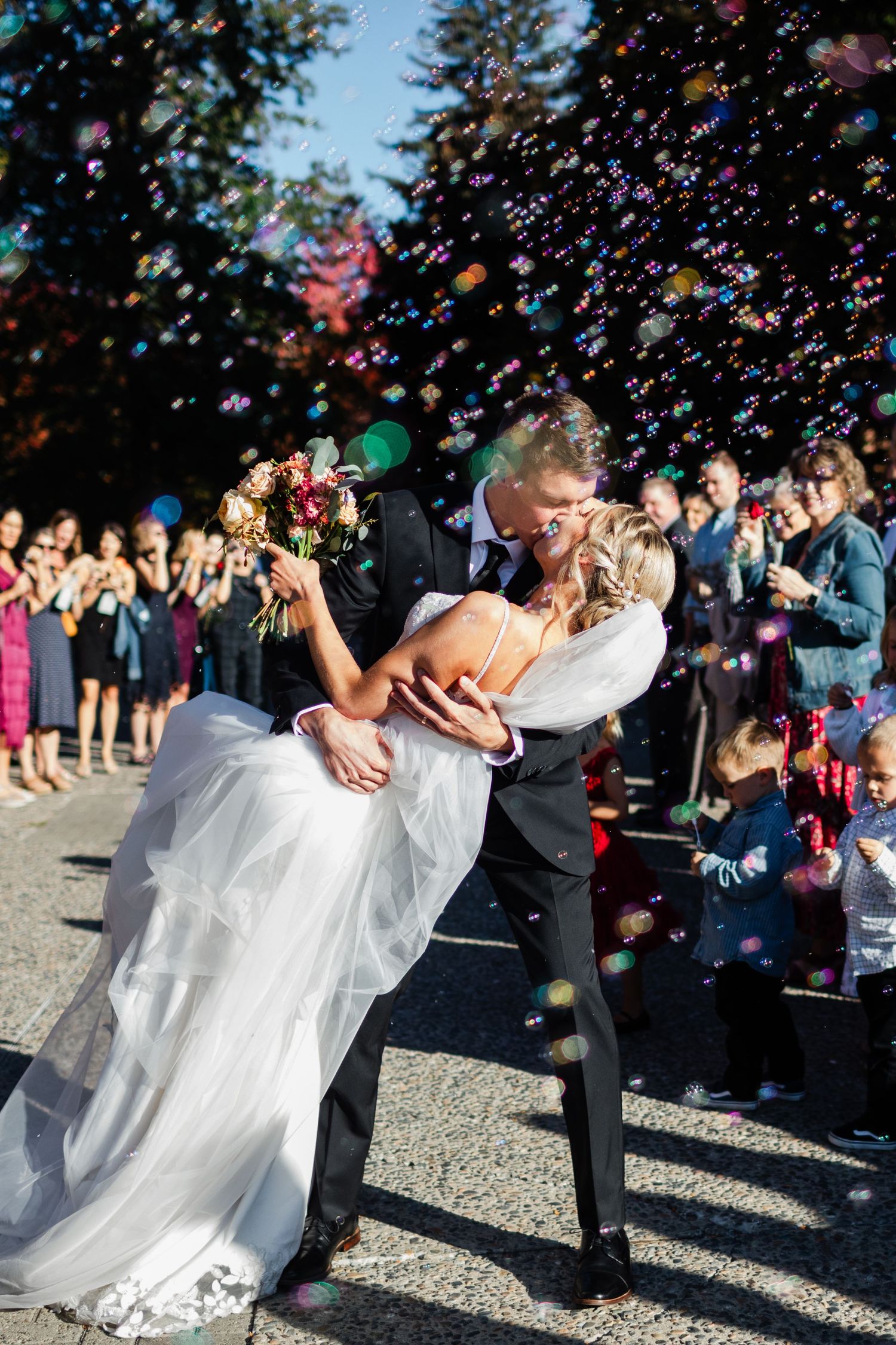 Groom dips bride for romantic kiss as guests celebrate with colorful confetti bubbles after wedding ceremony.