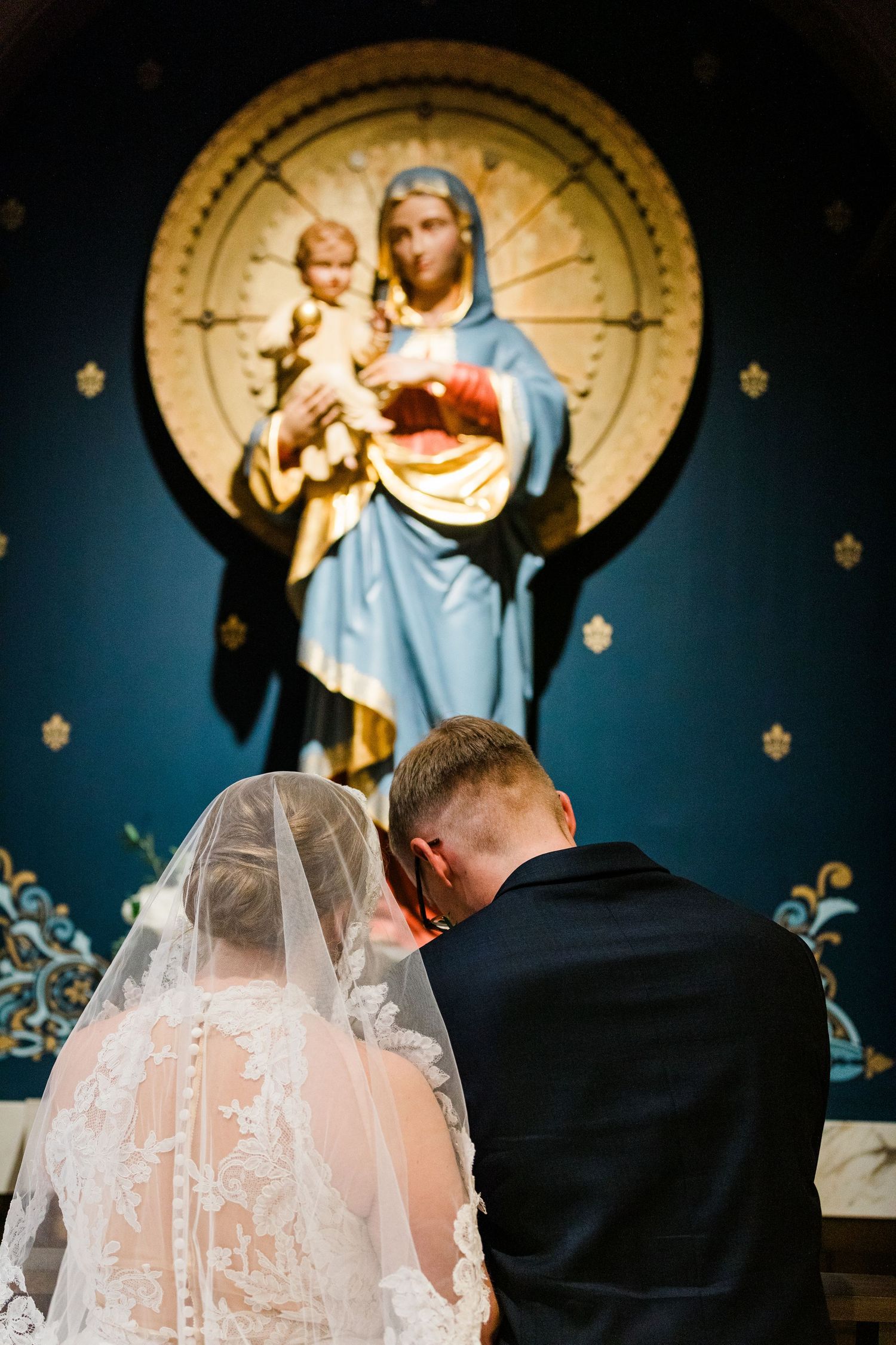Bride and groom bowing in prayer before a Virgin Mary statue during their Catholic wedding ceremony.