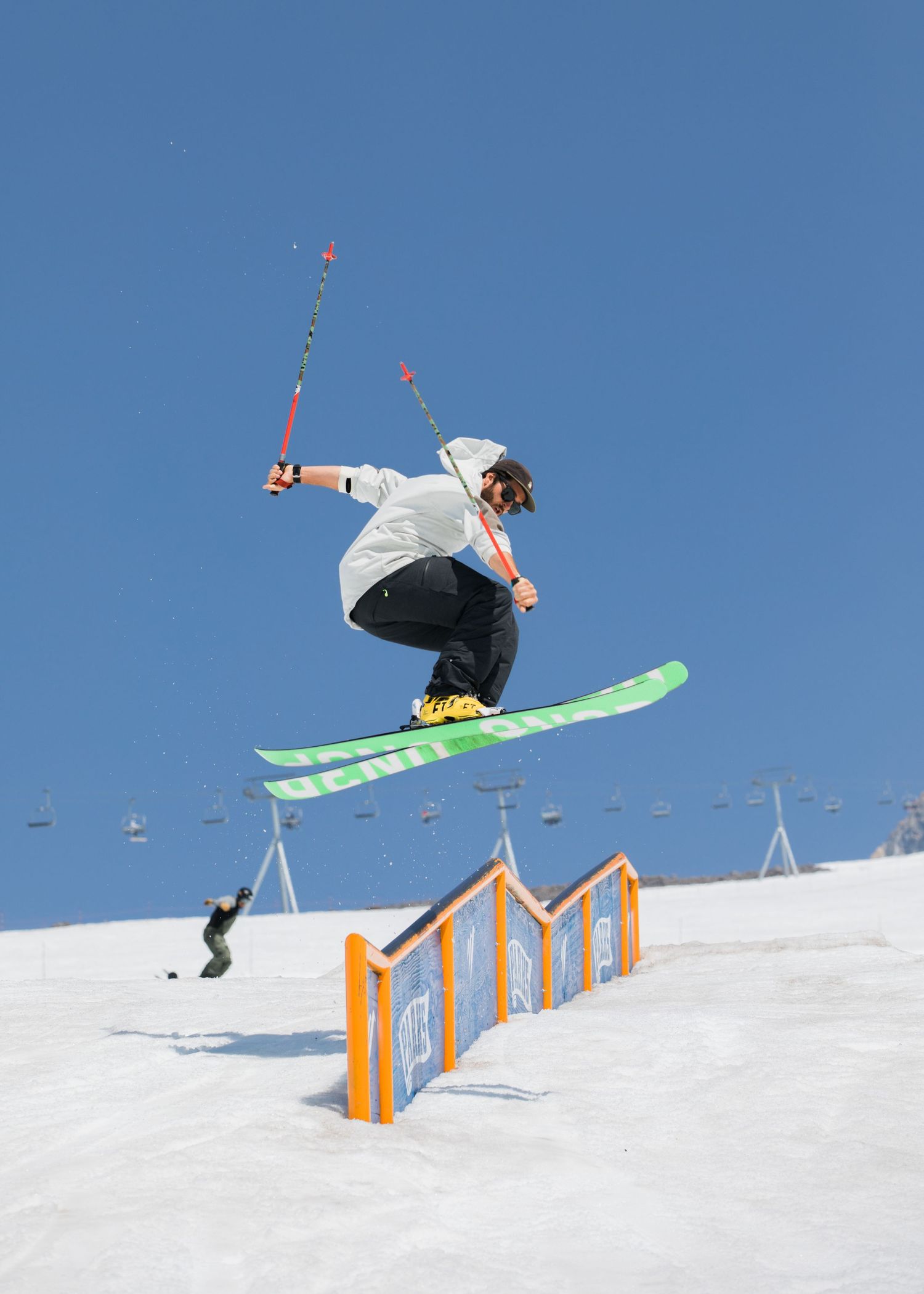 A skier performs a jump over an orange rail feature against a bright blue sky in a snow terrain park.