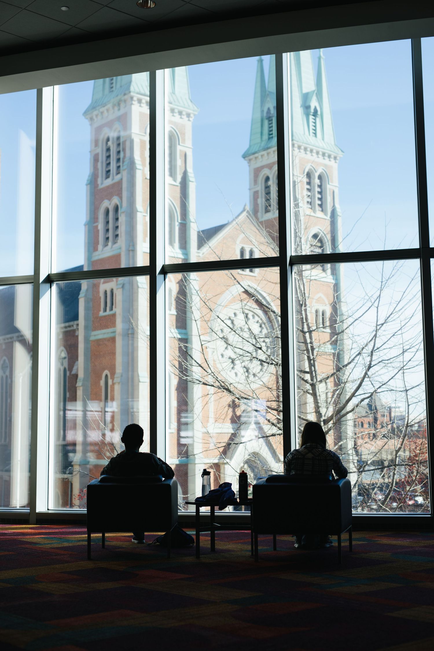 Two silhouettes enjoy a view of a grand cathedral through large windows in a quiet room.