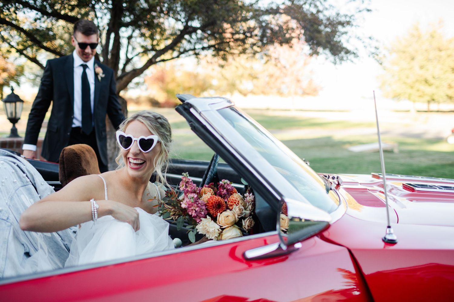 Bride in white dress and heart sunglasses sits in red convertible holding bouquet, groom in black suit stands behind.