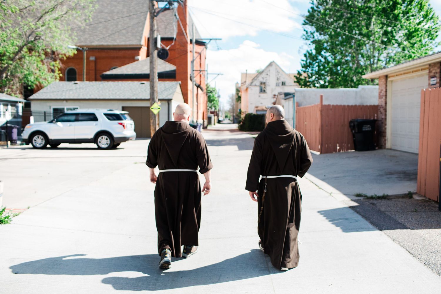 Two Catholic monks in black robes walk down a sunny suburban street lined with trees and parked cars.