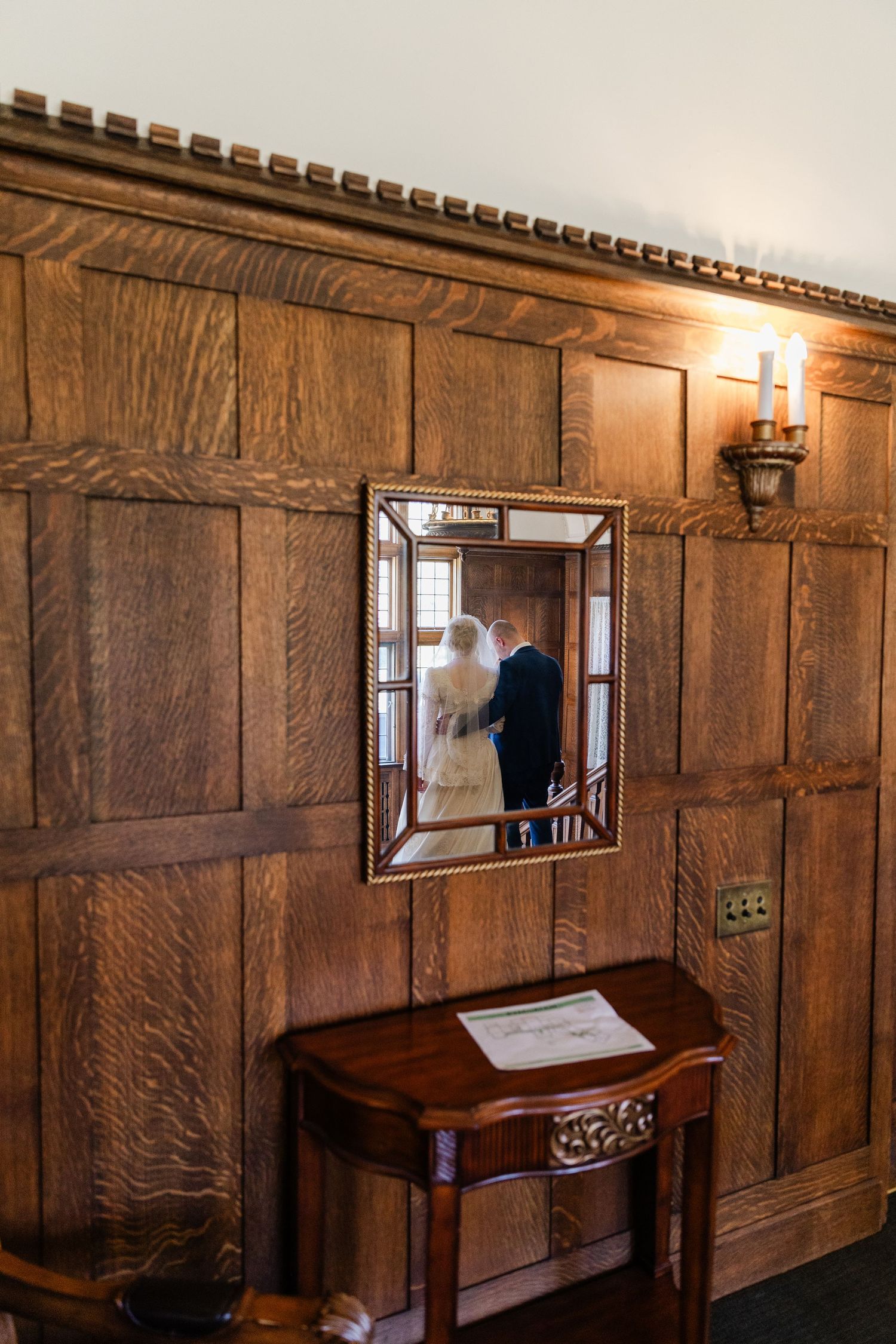 Bride and groom reflected in ornate gold mirror mounted on rich wood-paneled wall beside antique console table.