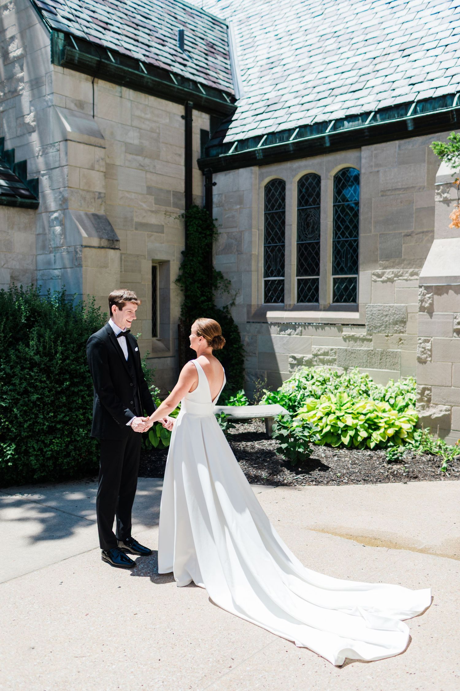 Bride in elegant white gown with long train holds hands with groom in black tuxedo outside stone church.