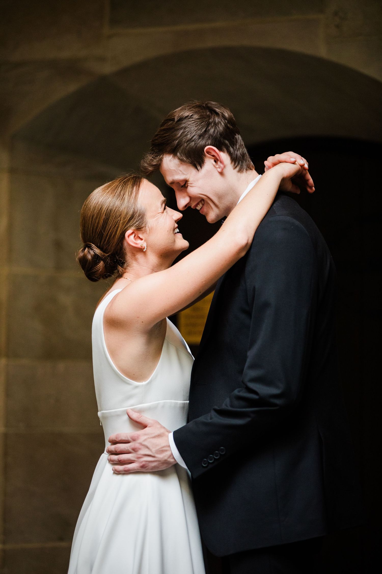 Happy newlyweds share romantic embrace smiling at each other against stone arch backdrop.