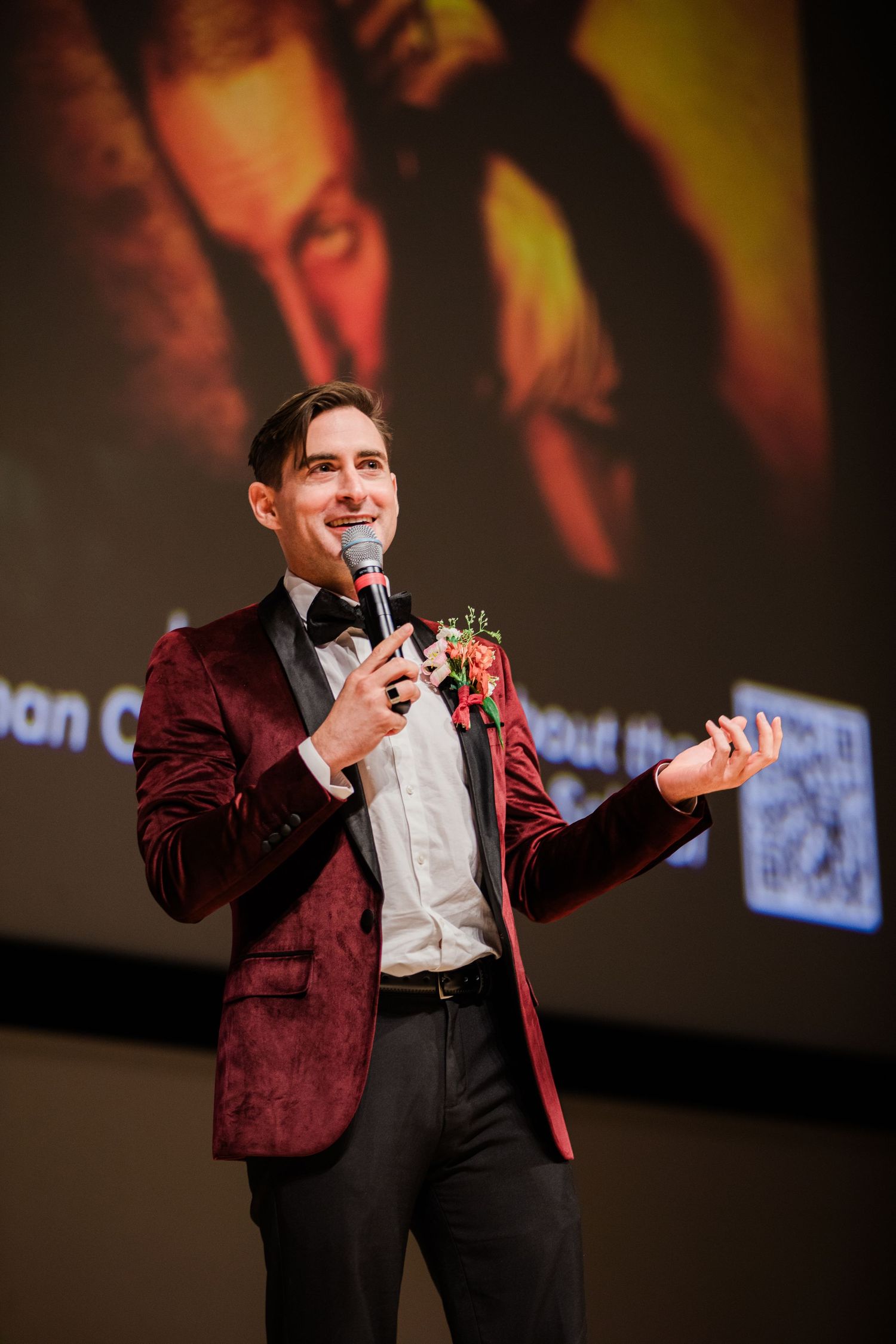 A performer in a burgundy velvet blazer speaks into a microphone on stage with dramatic lighting behind.