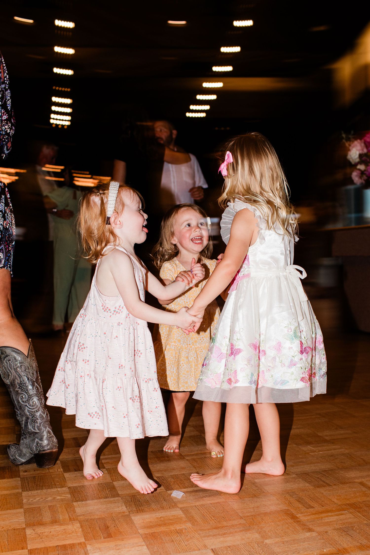 Young children in white dresses dancing and playing together on a wooden dance floor at a celebration.