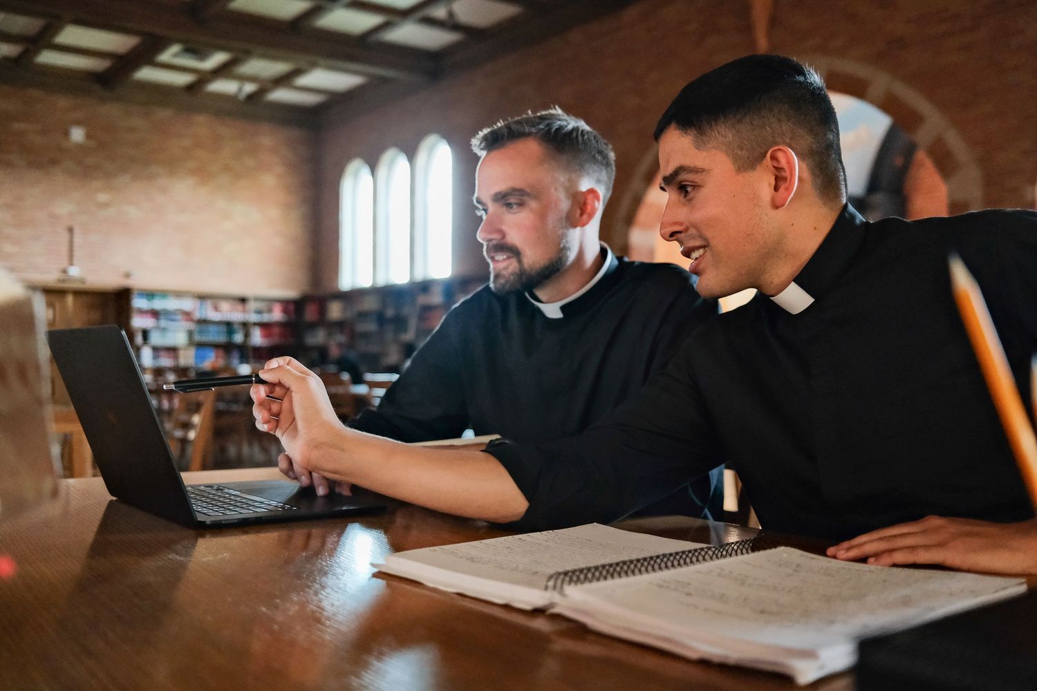 Two students study together at wooden tables in a library with warm lighting.
