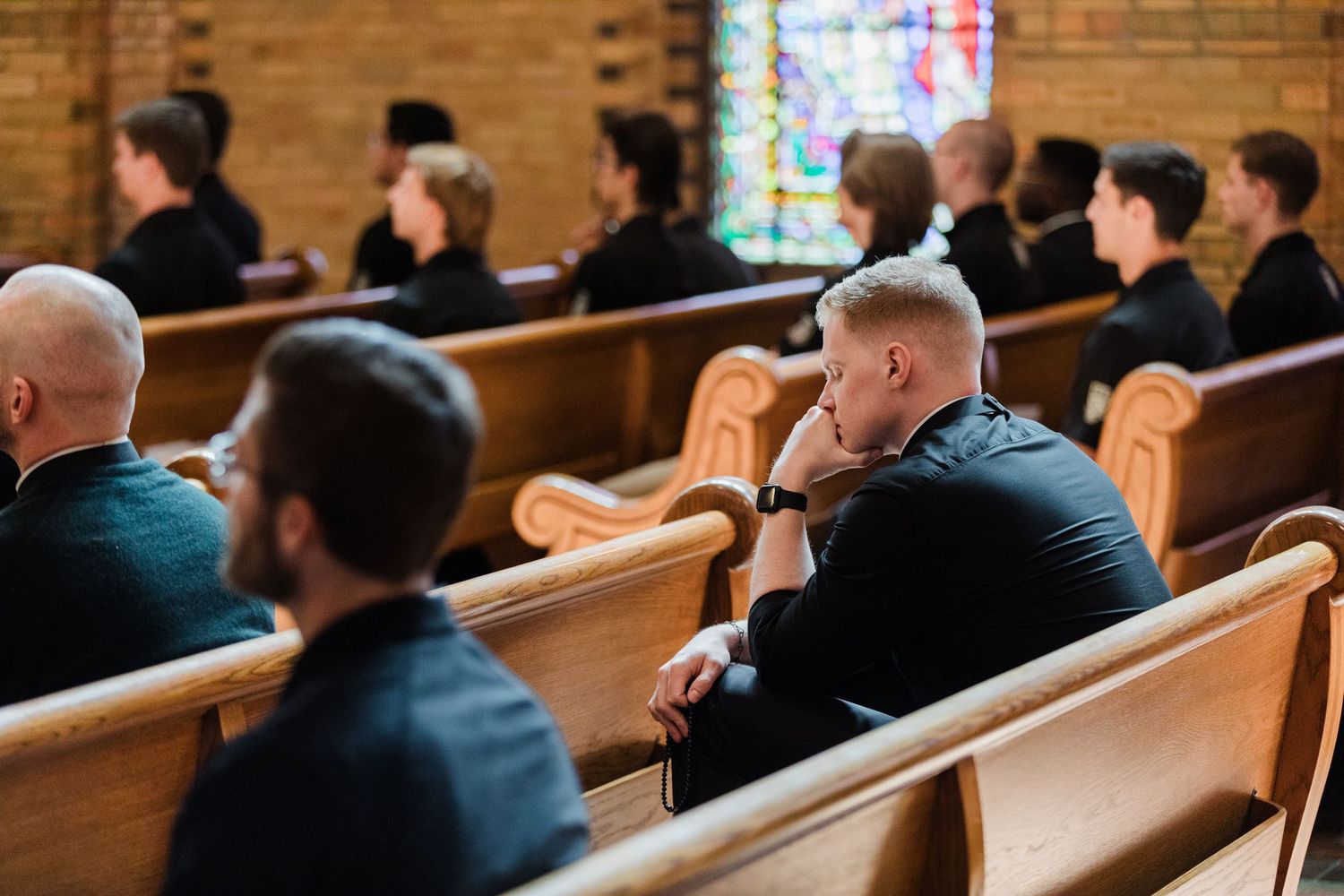 People seated in wooden church pews during a religious service with stained glass windows in background.
