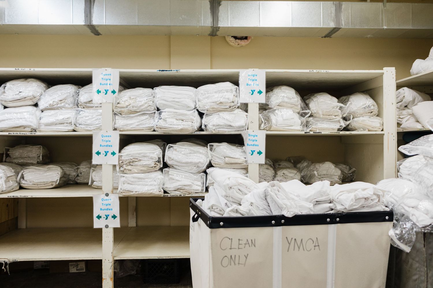 Shelves stocked with folded white linens and towels beside YMCA laundry carts labeled Clean Only.