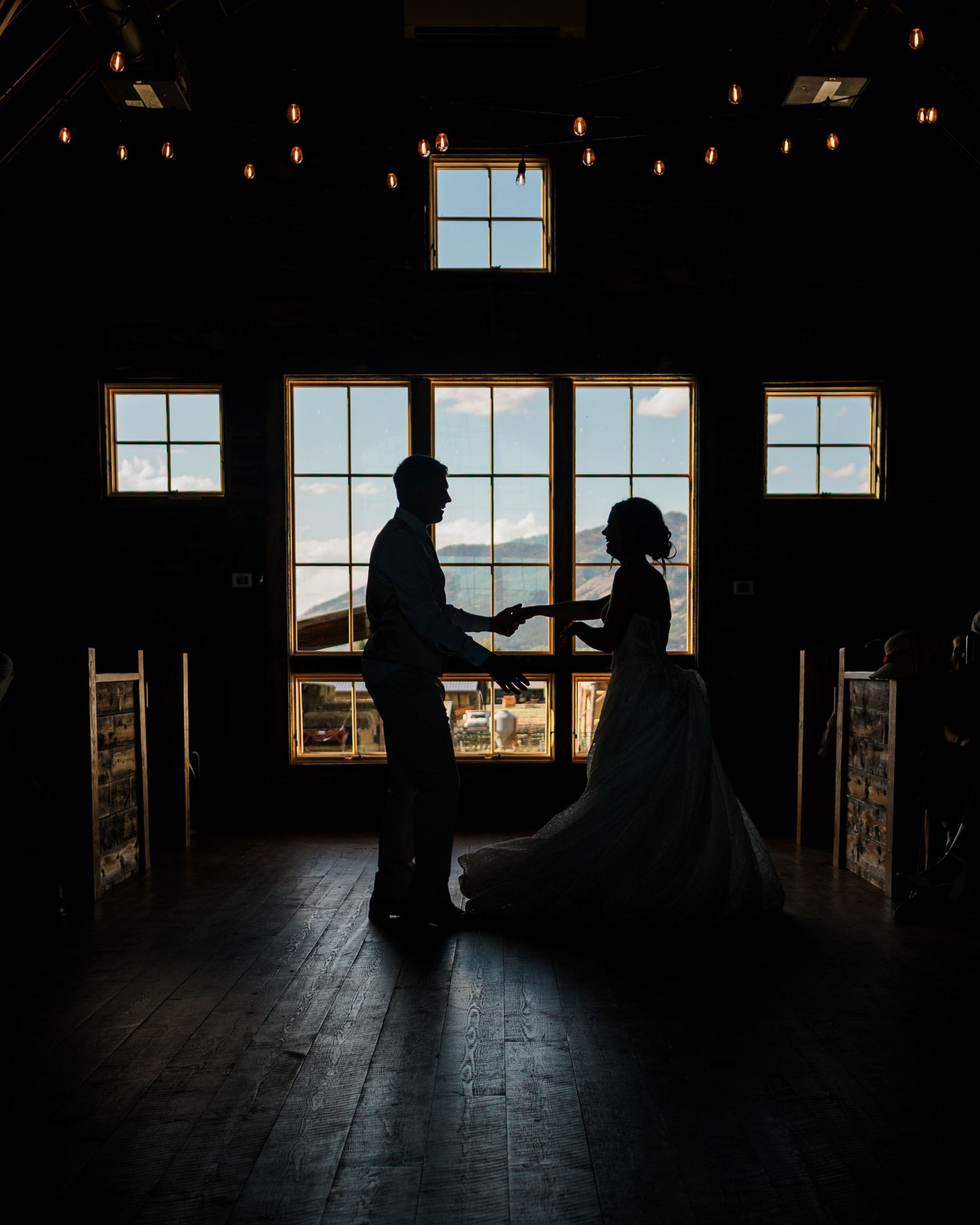 Silhouette of a bride and groom holding hands in a rustic barn venue with large windows and mountain views.