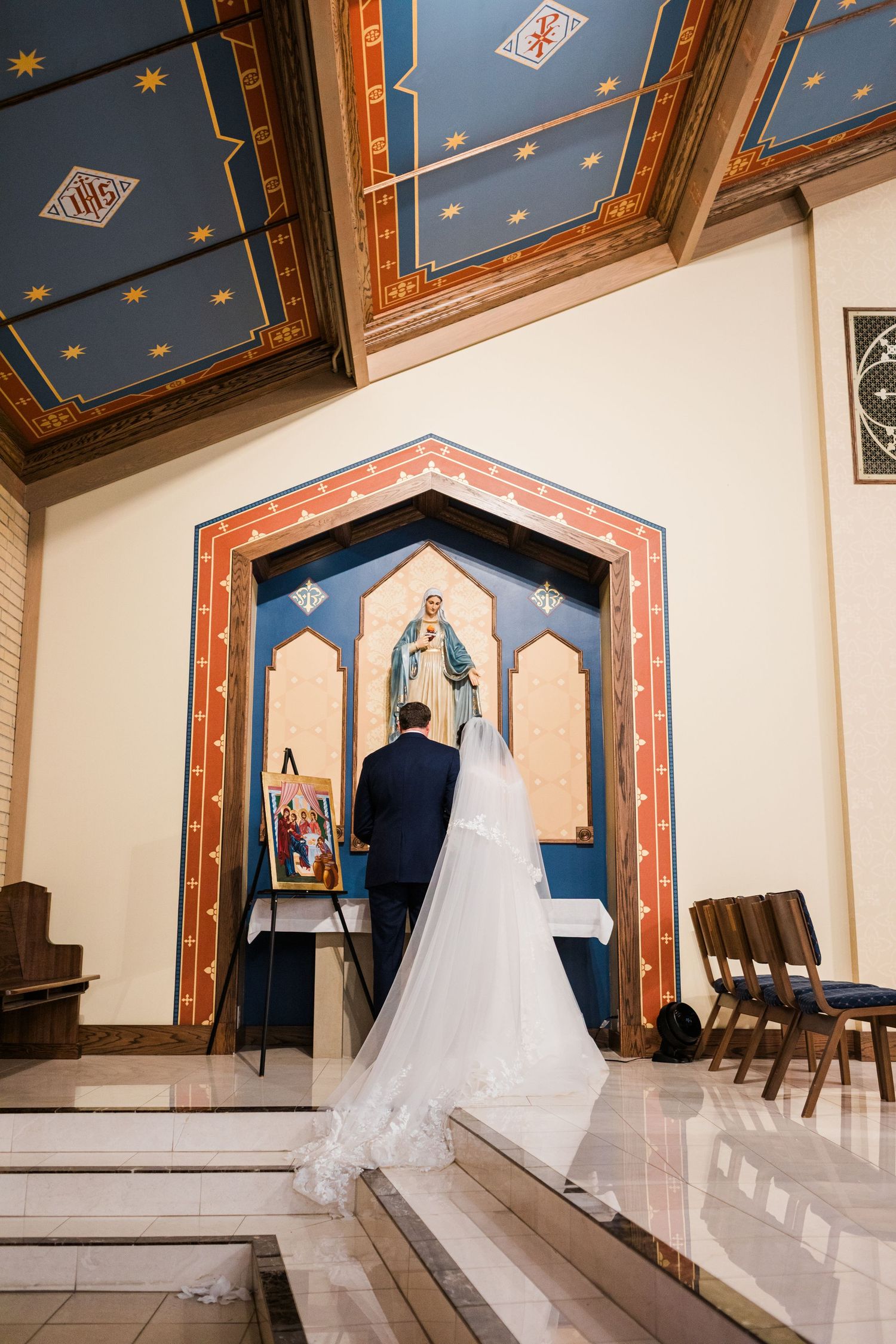 Bride and groom pray together before ornate religious shrine inside decorated Catholic church.