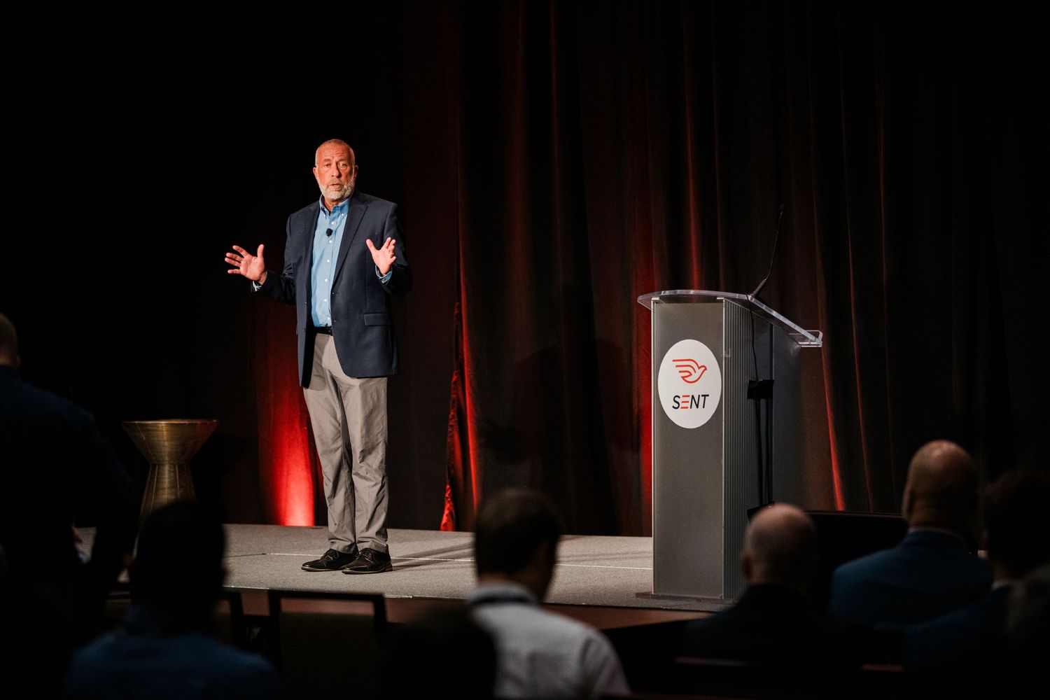 A speaker presents at a SUNY podium on a dark stage with red lighting during a conference event.