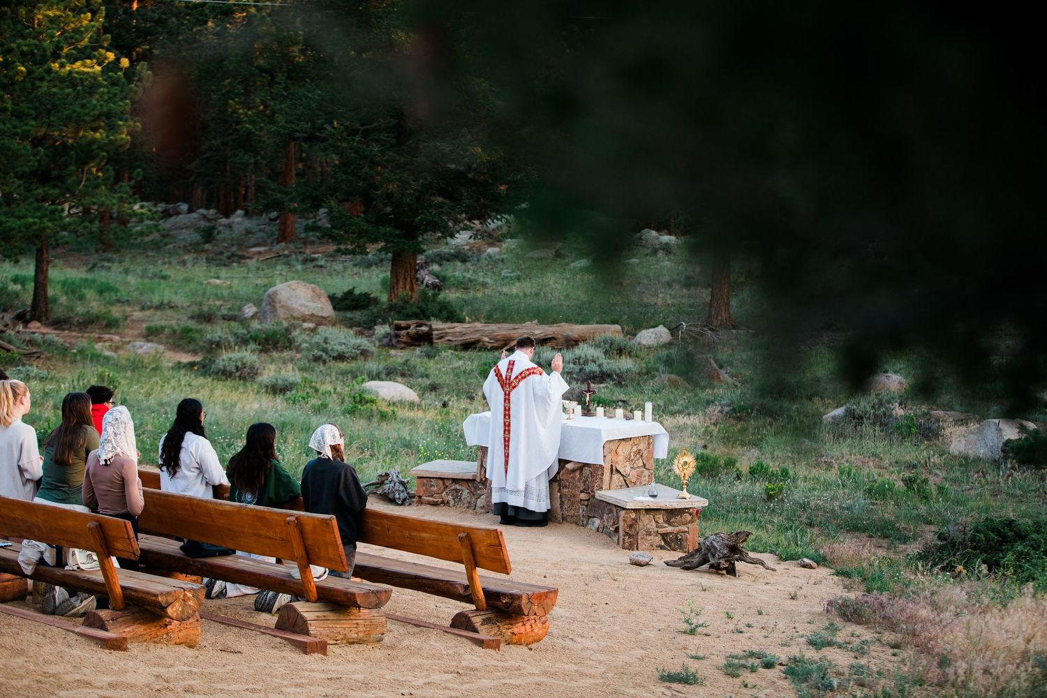 Outdoor Catholic Mass ceremony with priest at altar and congregation seated on wooden benches in a forest clearing.