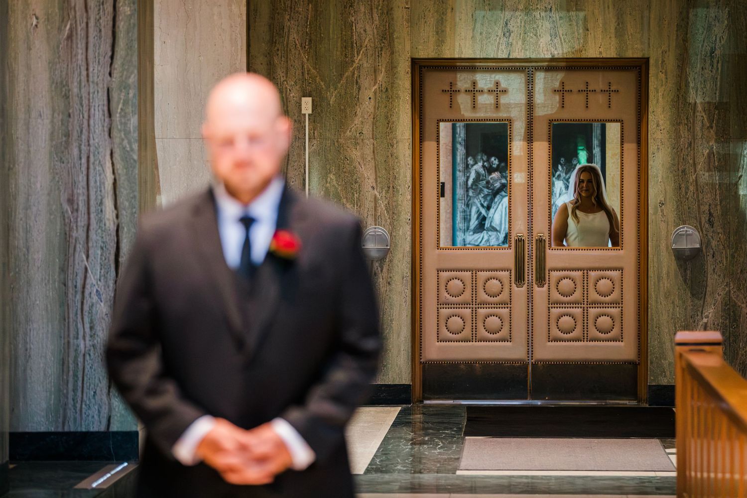 Groom in dark suit stands as bride approaches through ornate church doors in wedding first look.